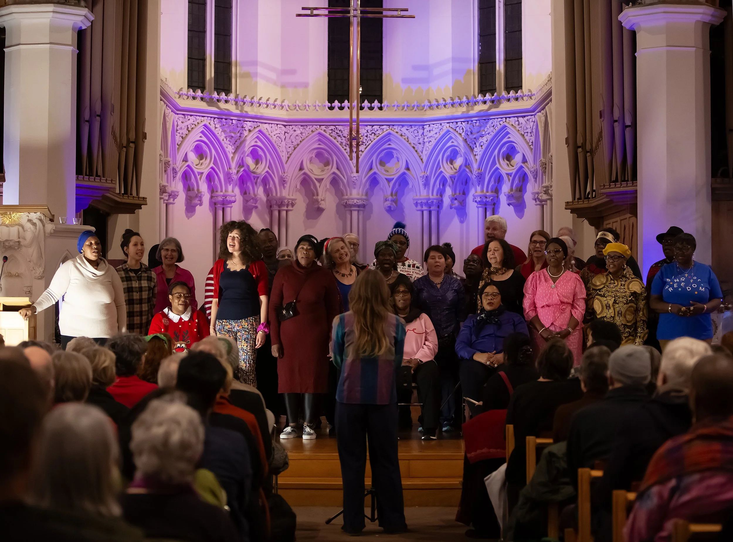 Group of diverse people standing on stage in a church with gothic architecture, purple lighting, and organ pipes, while a conductor leads them in a performance in front of an audience.