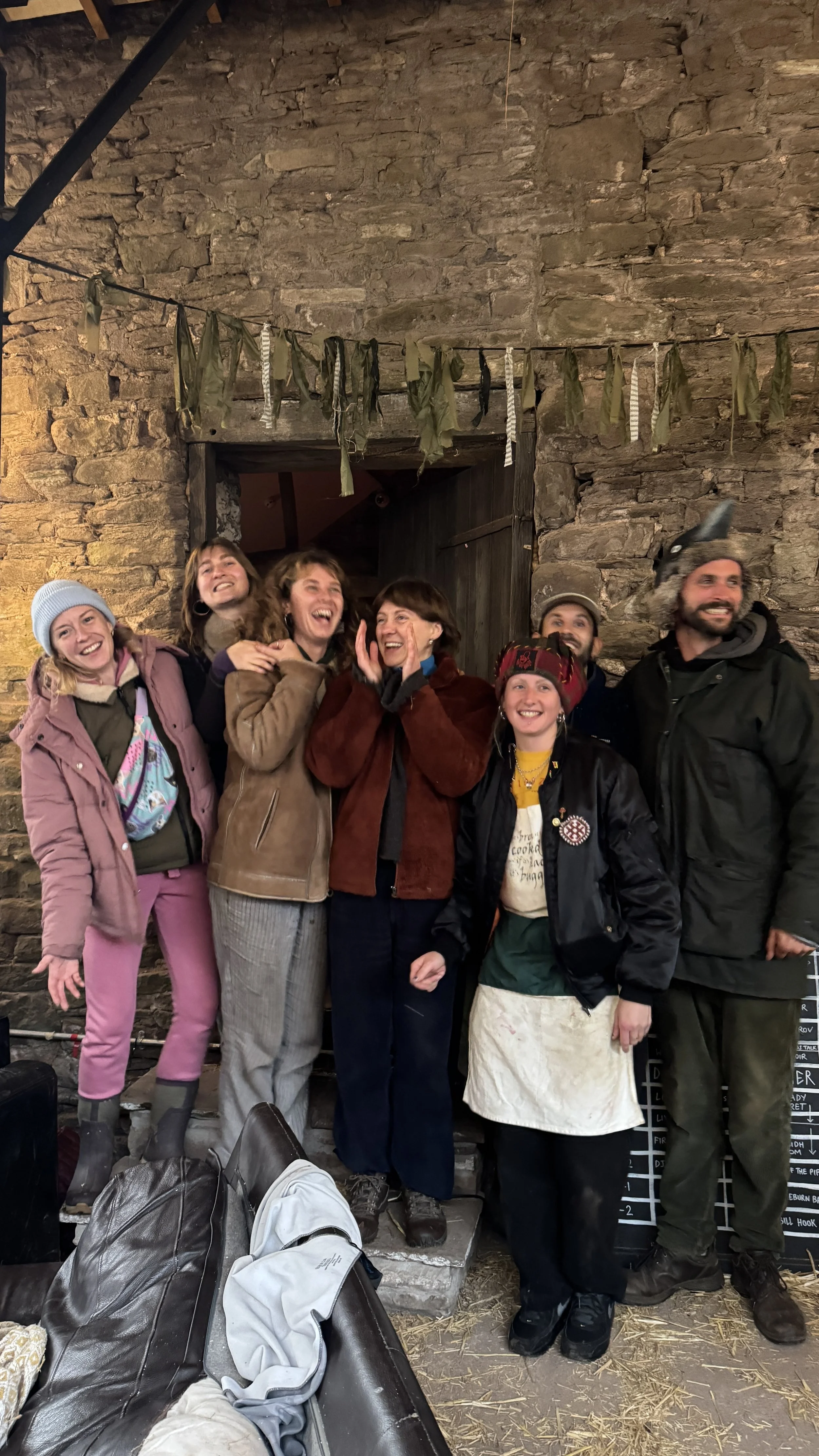 Group of seven people smiling and laughing inside a rustic stone building, some wearing outdoor clothing and one with a wolf hat, with a string of cloths hanging above.