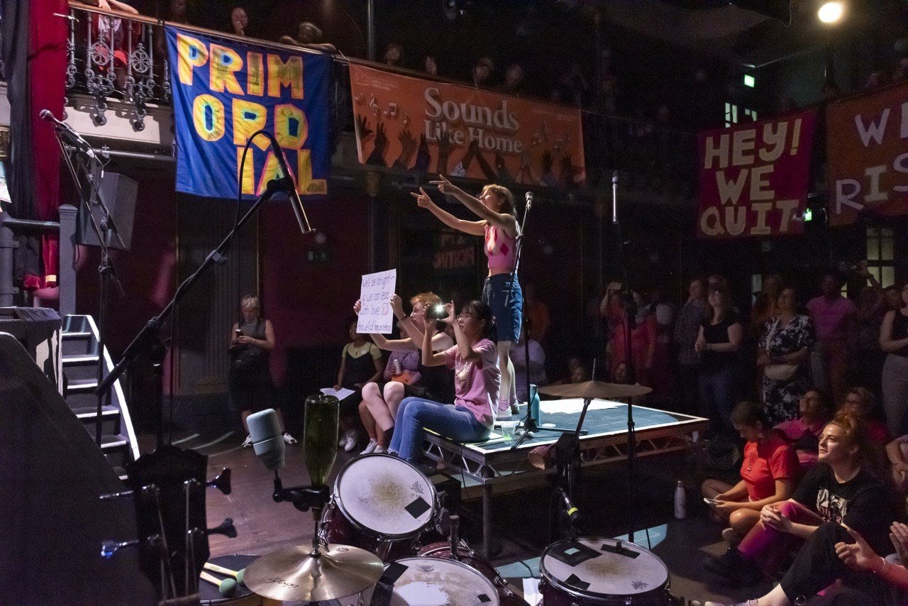 A woman on a platform points upward while another woman holds a sign, with a crowd watching at a concert or protest event. Banners read 'PRIM OPERAL' and 'HEY! WE QUIT' in the background.