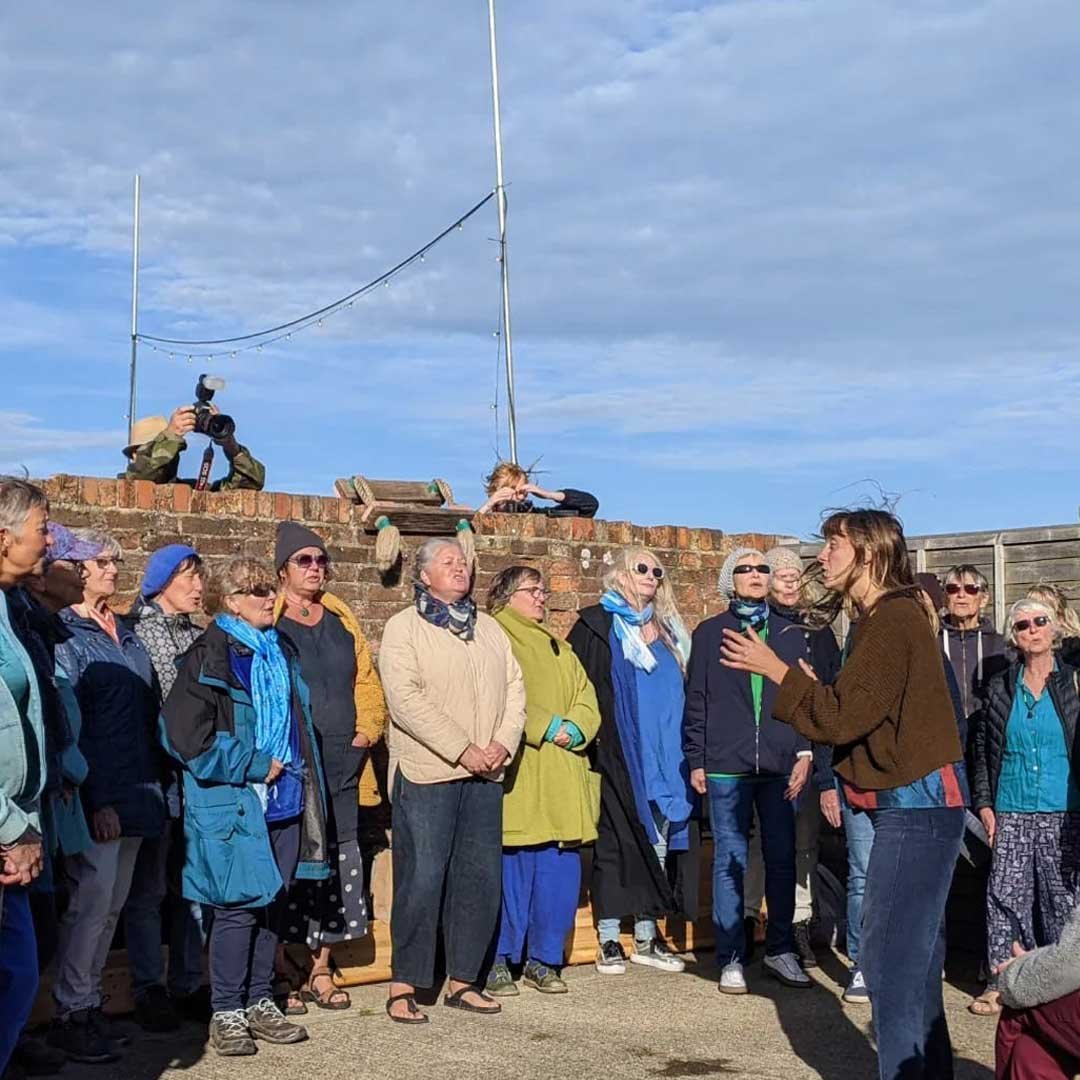 A tour guide speaking to a group of people outdoors, with a photographer taking pictures and a brick wall with poles and a cloudy sky in the background.