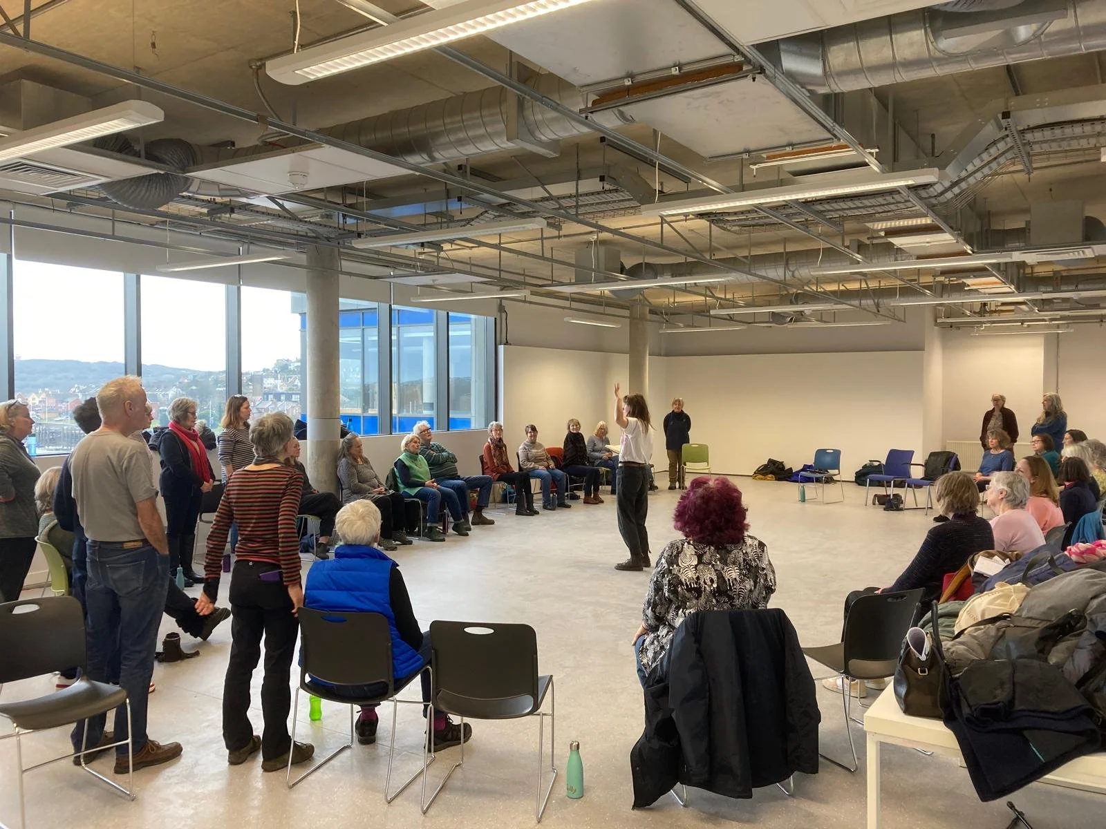 A person standing in front of a group of seated people in a spacious, modern room with large windows and an industrial ceiling.