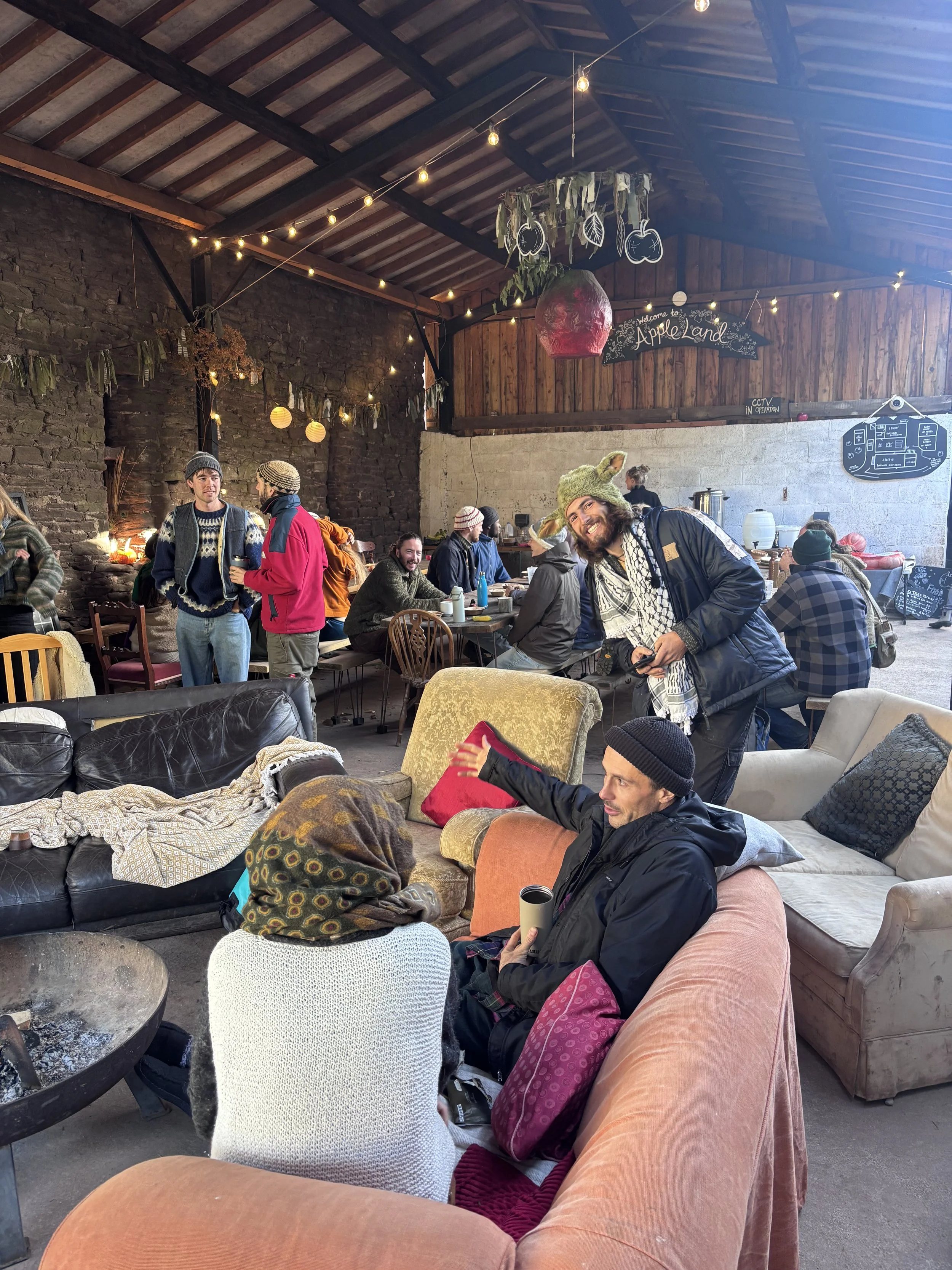 People gathered in a cozy, rustic indoor space decorated with string lights, hanging leaves, and pumpkins. Some are sitting on couches and armchairs, while others chat and stand in the background.