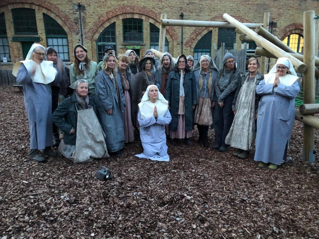 A group of women dressed as nuns and period clothing poses together outdoors on a gravel surface, with some standing and one kneeling, in front of a brick building with arched windows and a wooden playground structure.