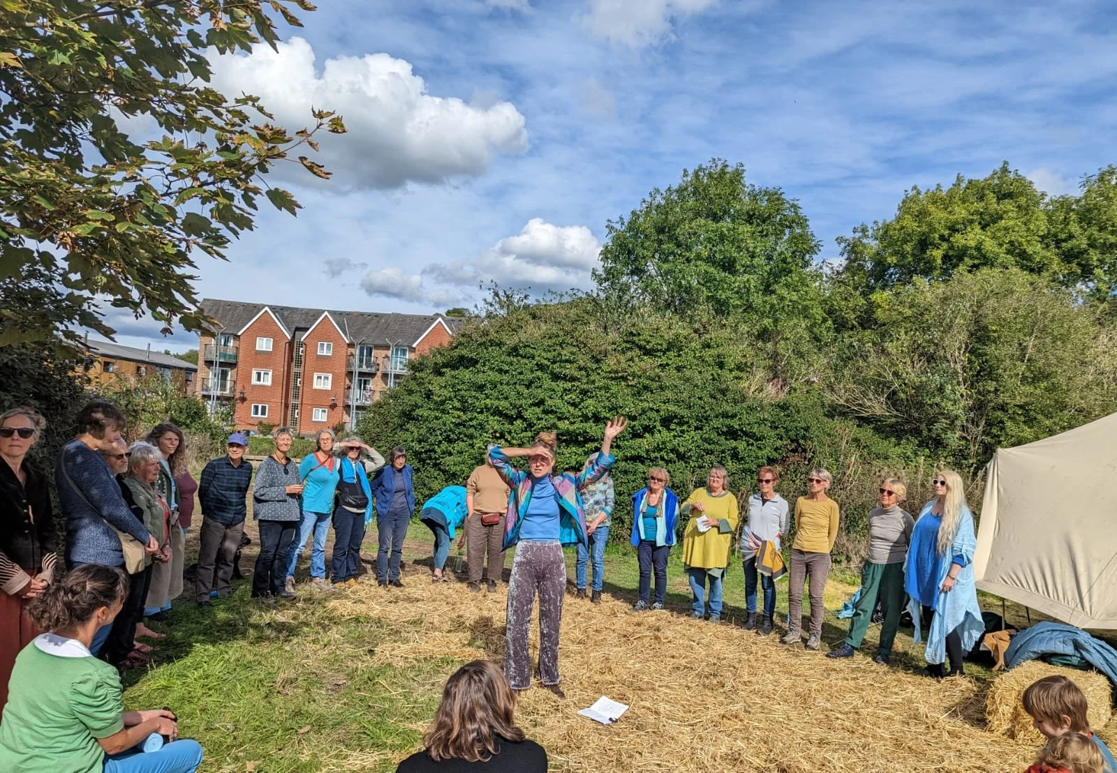 Group of people outdoors in a grassy area with hay bales, listening to a woman speaking with her hands raised. Trees and a multi-story brick building are in the background, under a partly cloudy sky.
