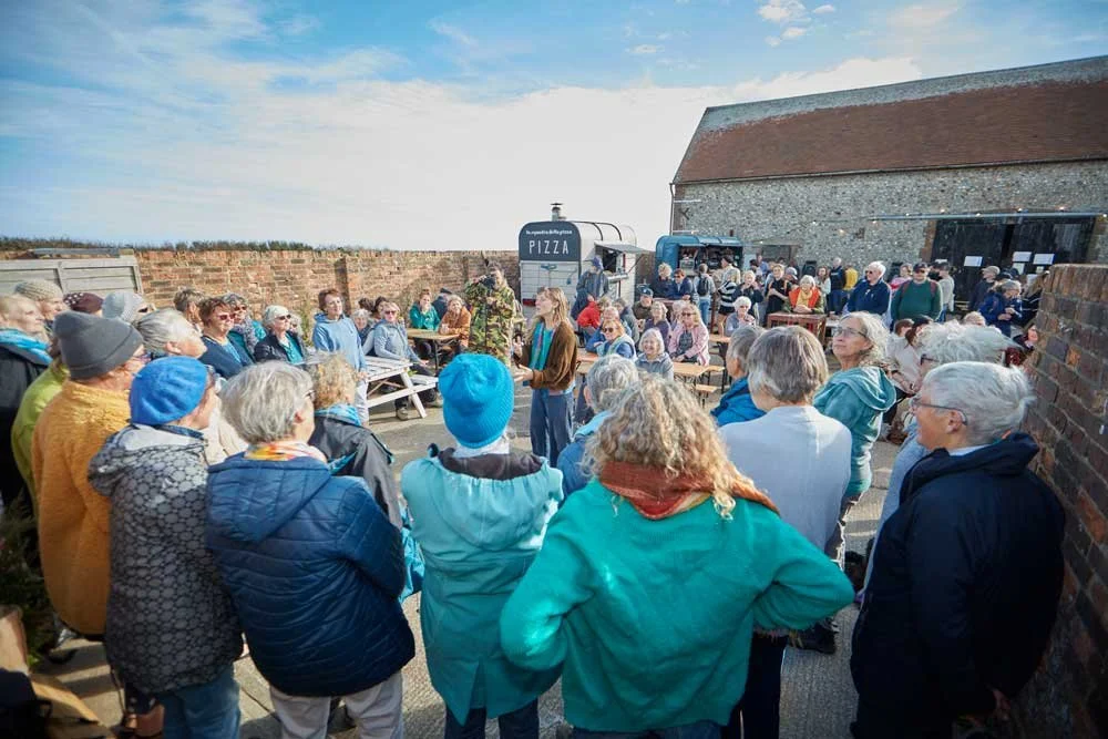 A large group of people gathered outdoors at a community event, listening to a woman speaking with a food truck and a sign that says 'PIZZA' in the background.