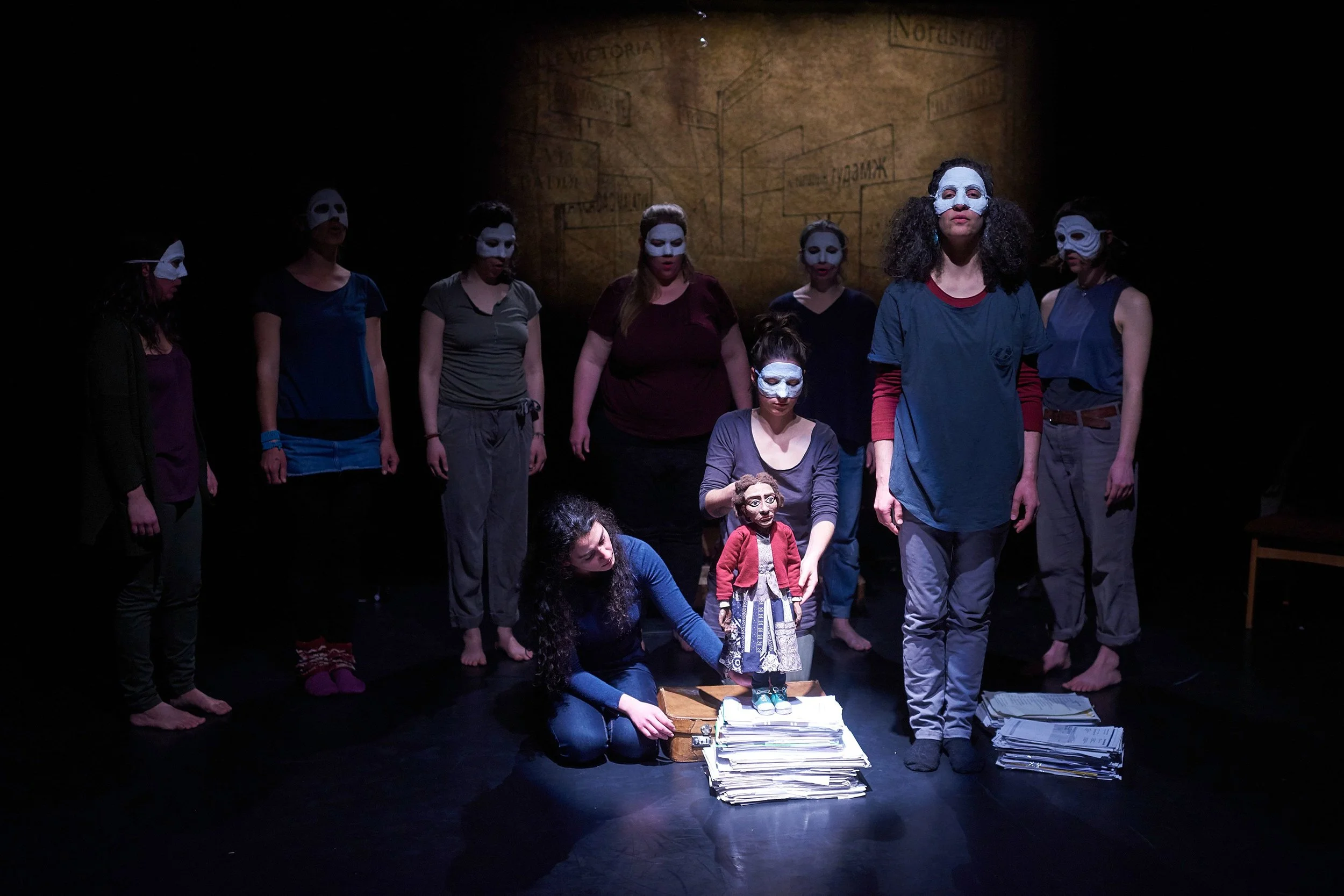 Group of people wearing masks standing on stage, with one person kneeling and handling a puppet and papers, in a theatrical setting.