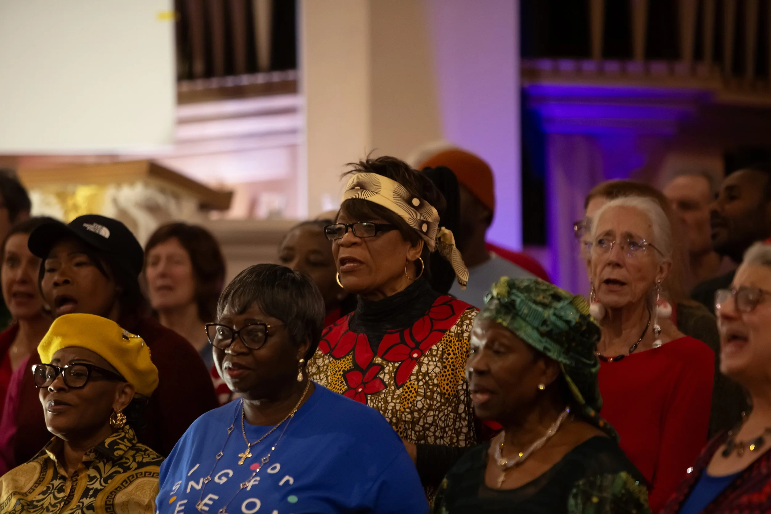 Group of diverse women singing at an indoor event, some wearing colorful headscarves and glasses, with a background of warm lighting and purple accent lights.