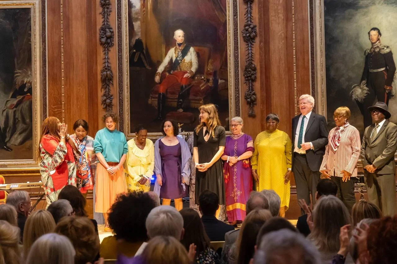 A diverse group of people stands on a stage in front of a large ornate painting, facing an audience. The group appears to be at an event or celebration.