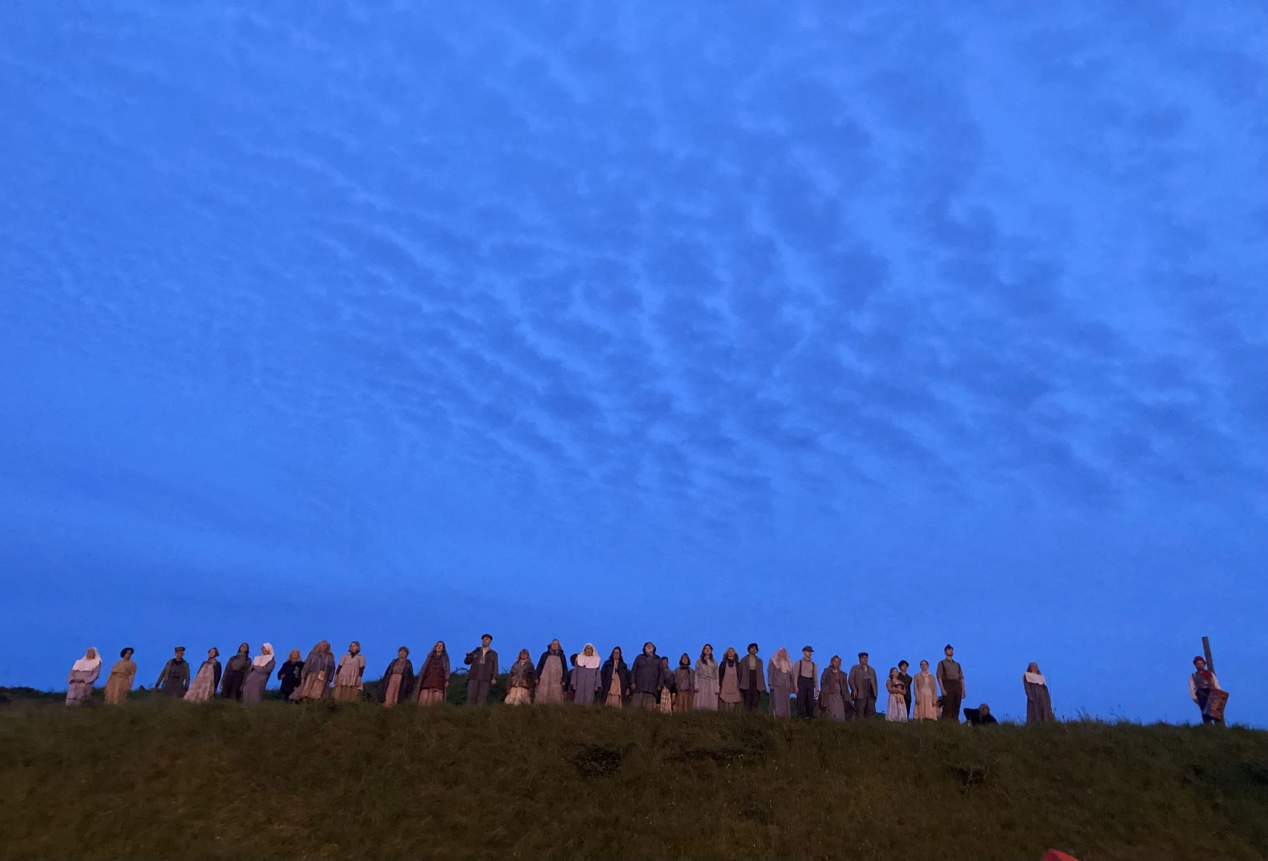 A line of people dressed in historical clothing standing on a grassy hill under a blue sky with neural cloud formations.