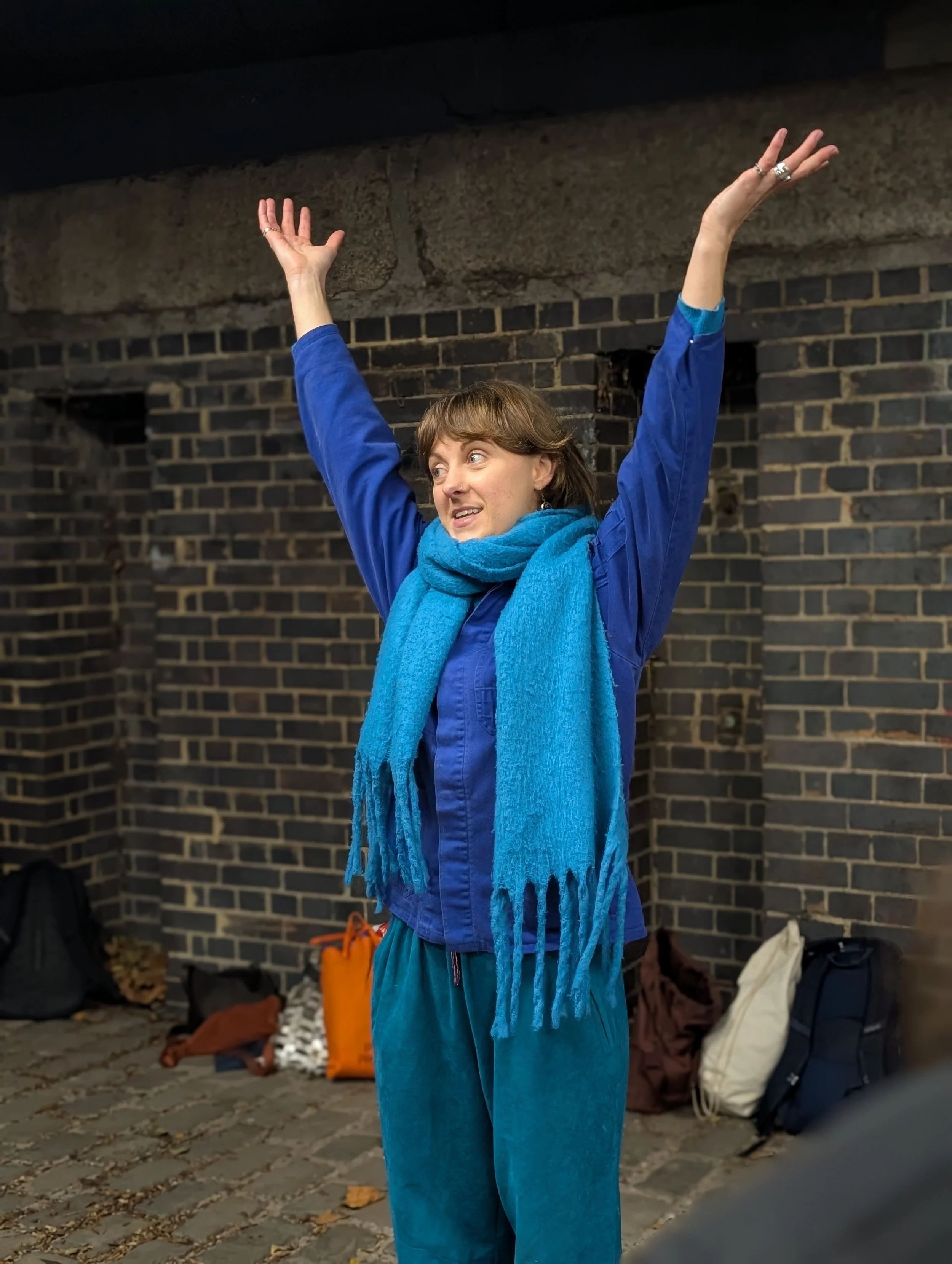 A woman wearing a blue jacket and a blue scarf, standing outside with her arms raised, smiling. She is in front of a brick wall with backpacks and bags on the ground.