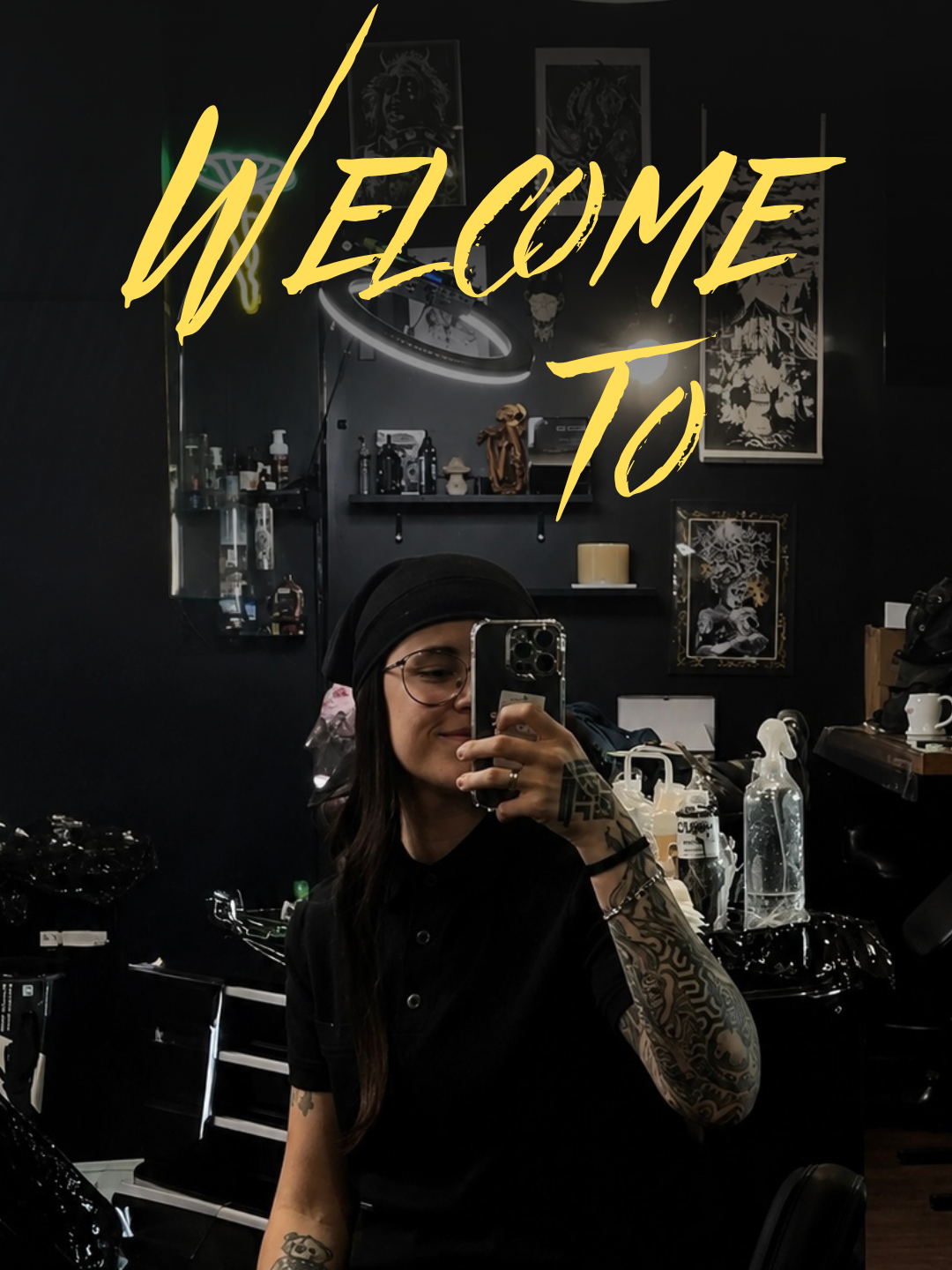 Copito taking a mirror selfie in the dark tattoo studio, with a yellow WELCOME TO graphic across the top and framed artwork behind