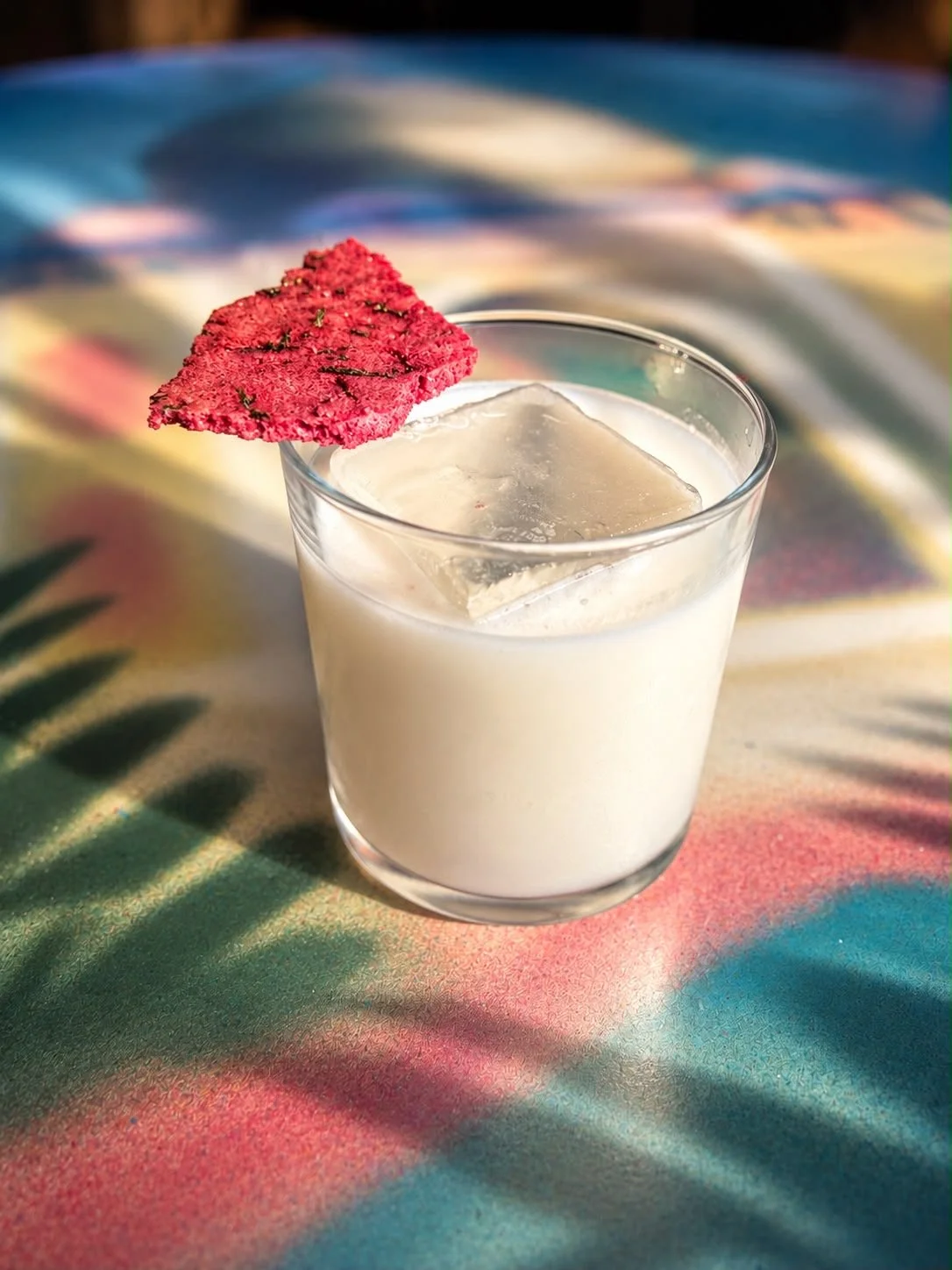 Creamy white cocktail over a large ice cube with a red crisp garnish on a colourful table