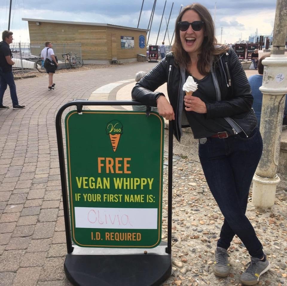 OffersOn founder Olivia Gray laughing on Brighton seafront holding an ice cream next to a sign offering a free vegan whippy to anyone named Olivia