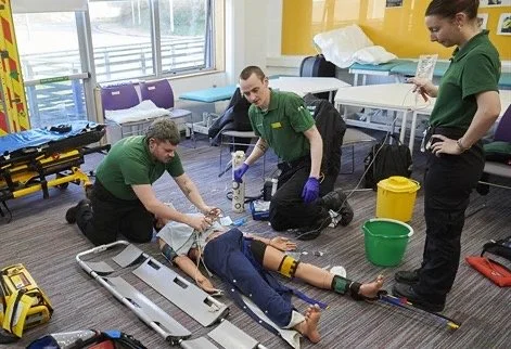 Three University of Brighton paramedic science students in green uniforms practising emergency response around a mannequin in a training room