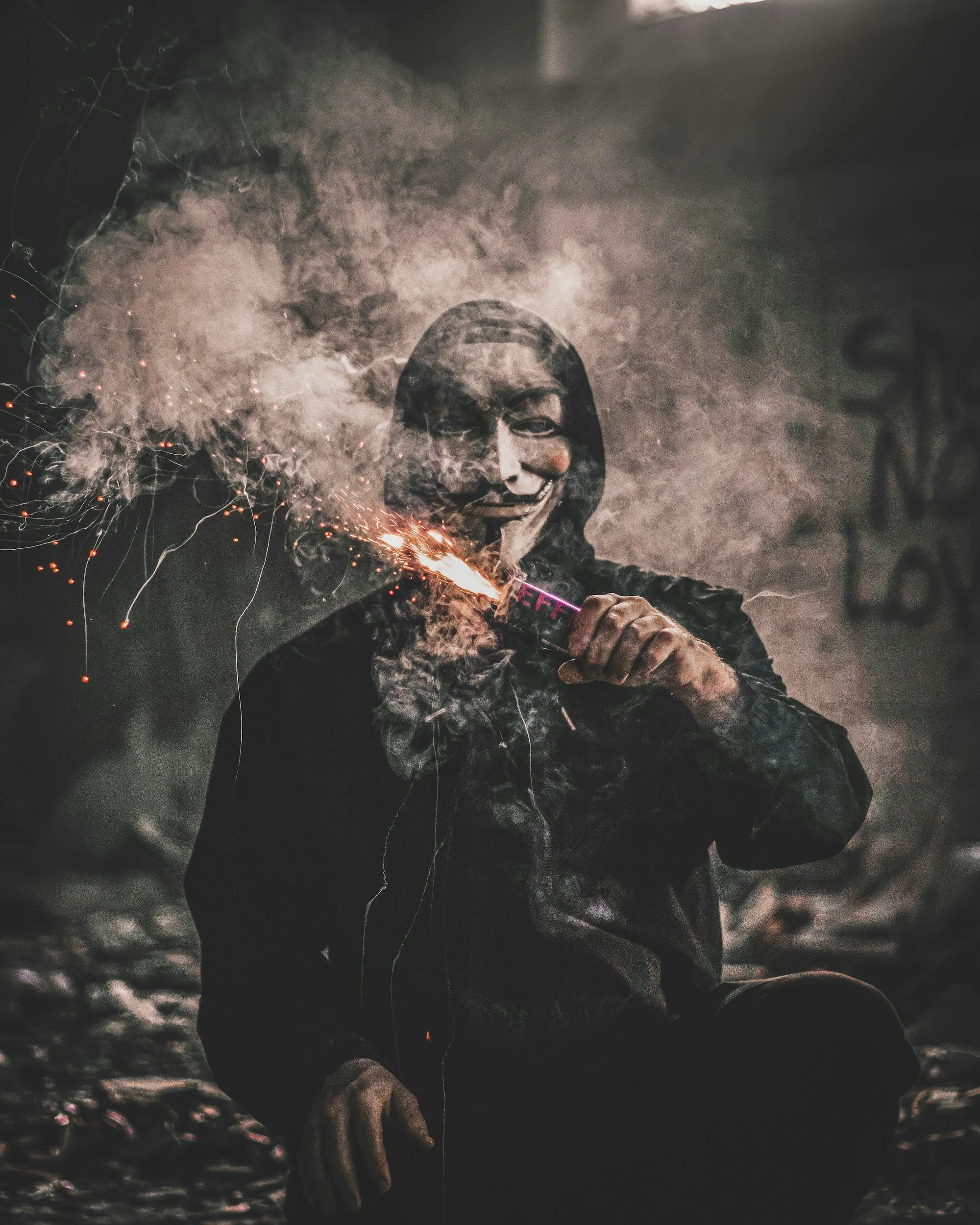 Person wearing a Guy Fawkes mask holding a firework with sparks and smoke around.