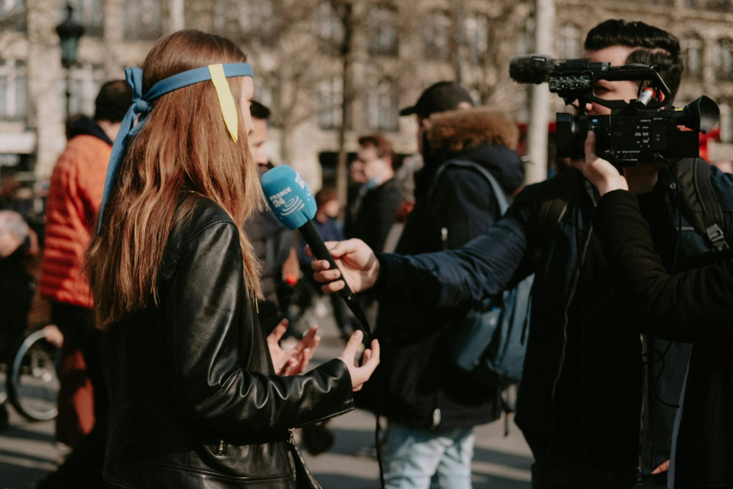A woman being interviewed outdoors by a person holding a microphone labeled 'France 24'. A cameraman is filming the interview, and there are people and trees in the background.