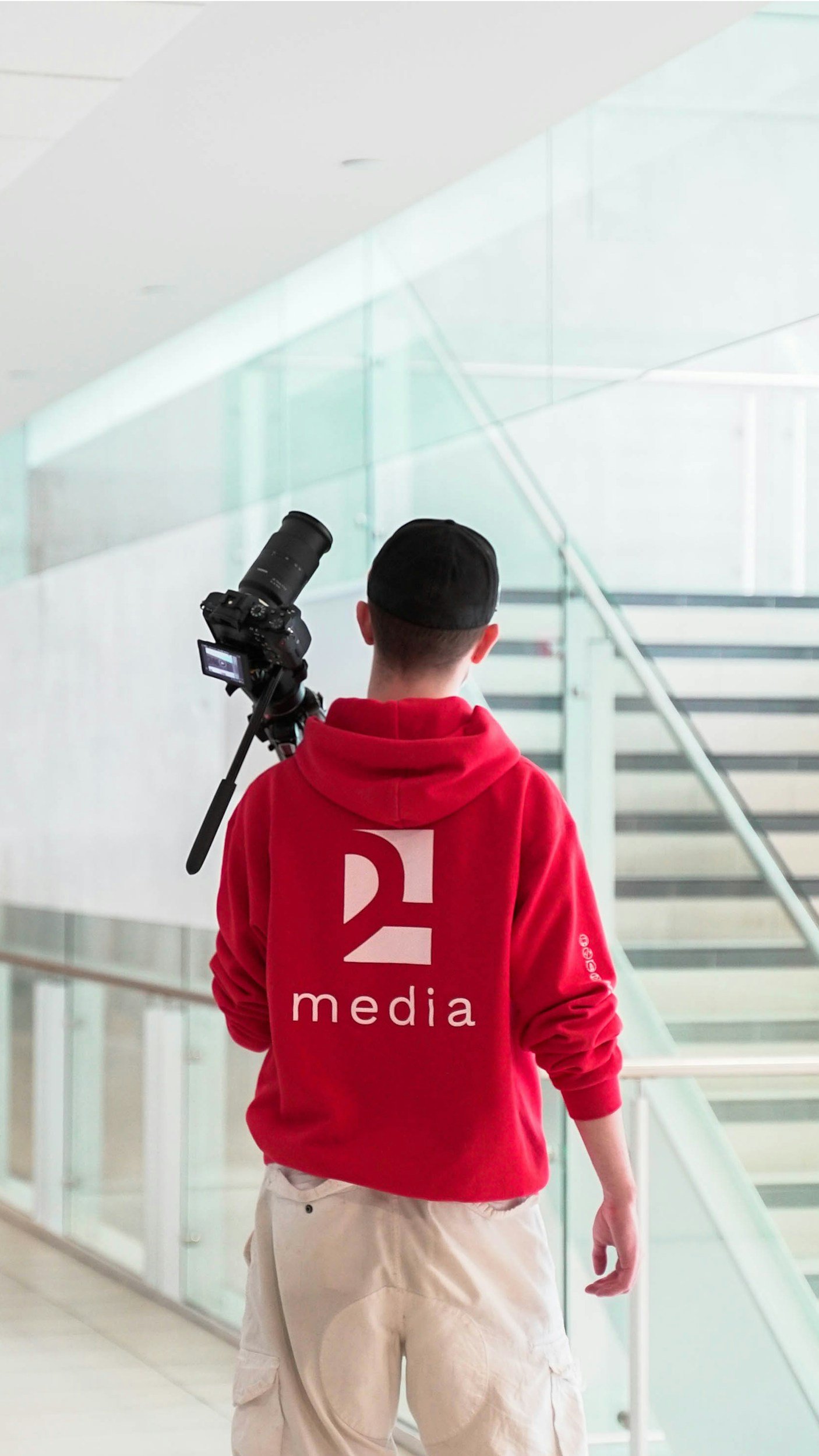 A person seen from behind, wearing a red hoodie with the logo '2 media' on the back, holding a camera, and walking up an indoor staircase in a modern building with glass railings.