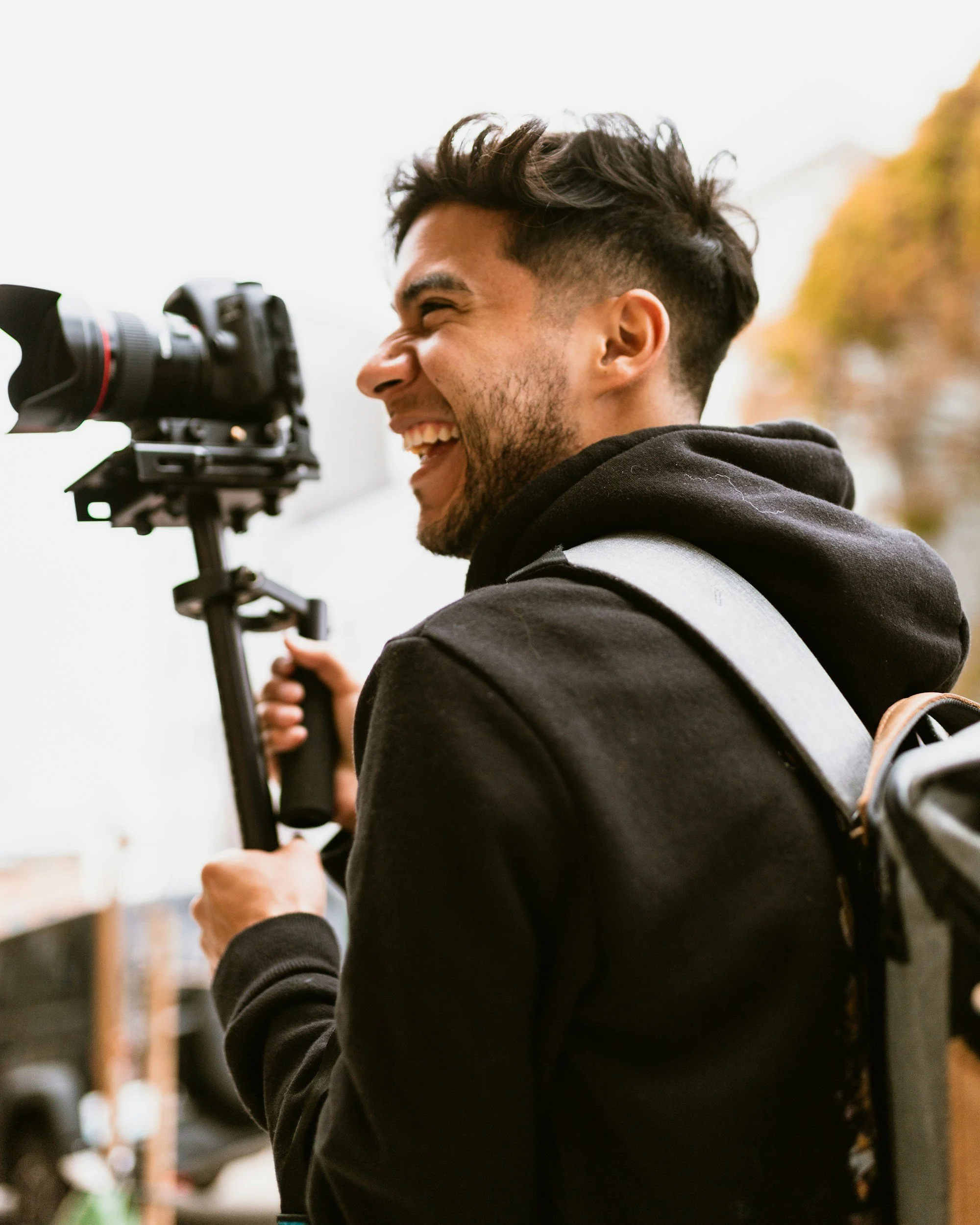 A young man with dark hair and a beard, smiling, holding a camera on a tripod outdoors during daytime, with trees in the background.
