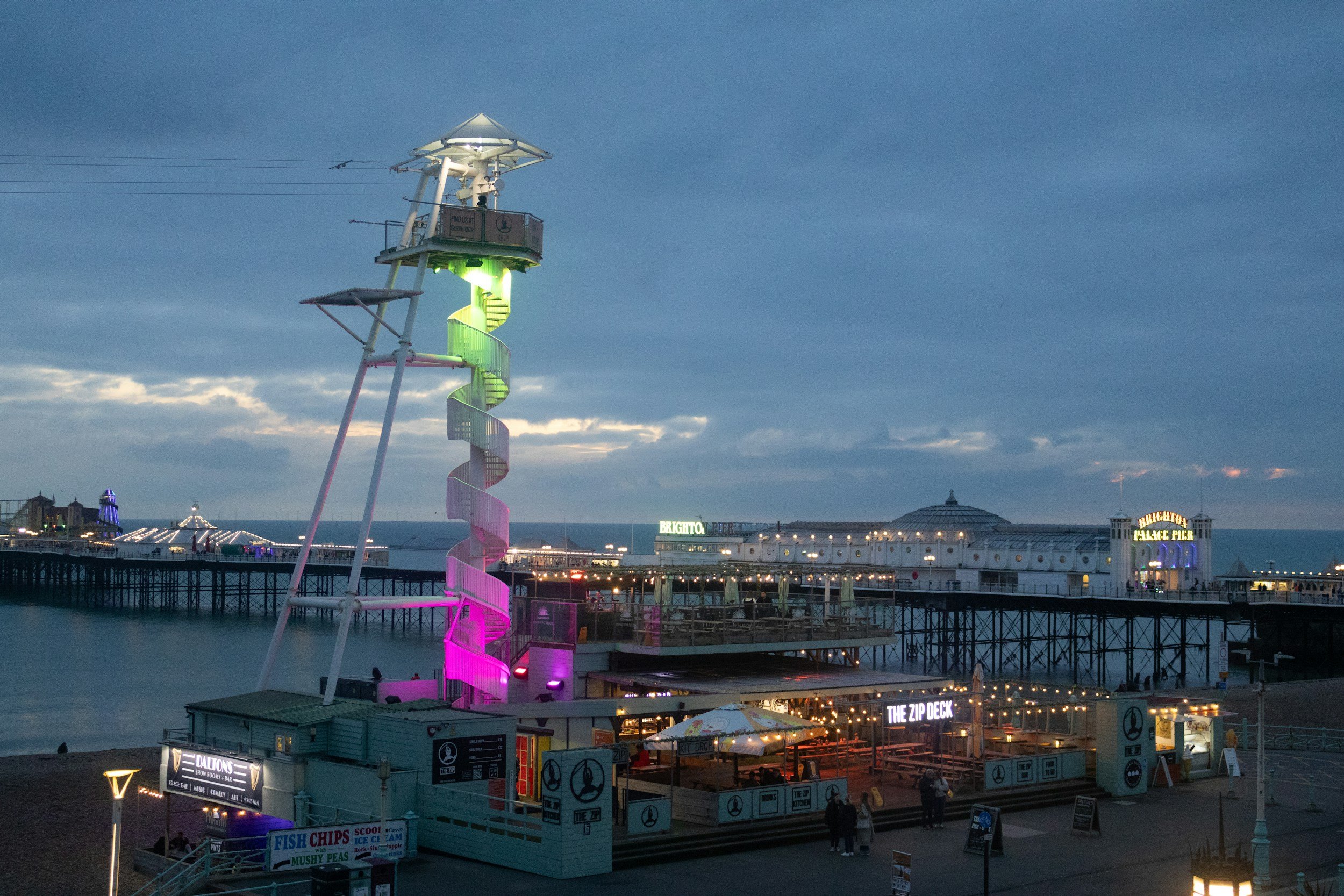 The Brighton Zip tower lit up at dusk with Palace Pier and the seafront behind