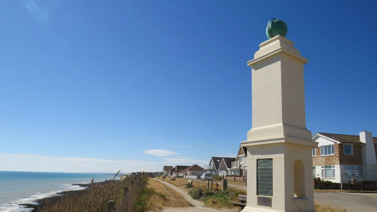 The Greenwich Meridian monument at Peacehaven, a pale stone column on the cliff top beside the sea under a blue sky