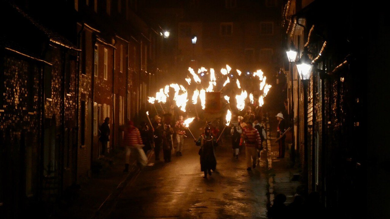 Torch-bearing bonfire society members walking along a dark Lewes street at night, with flaming torches lighting the shopfronts