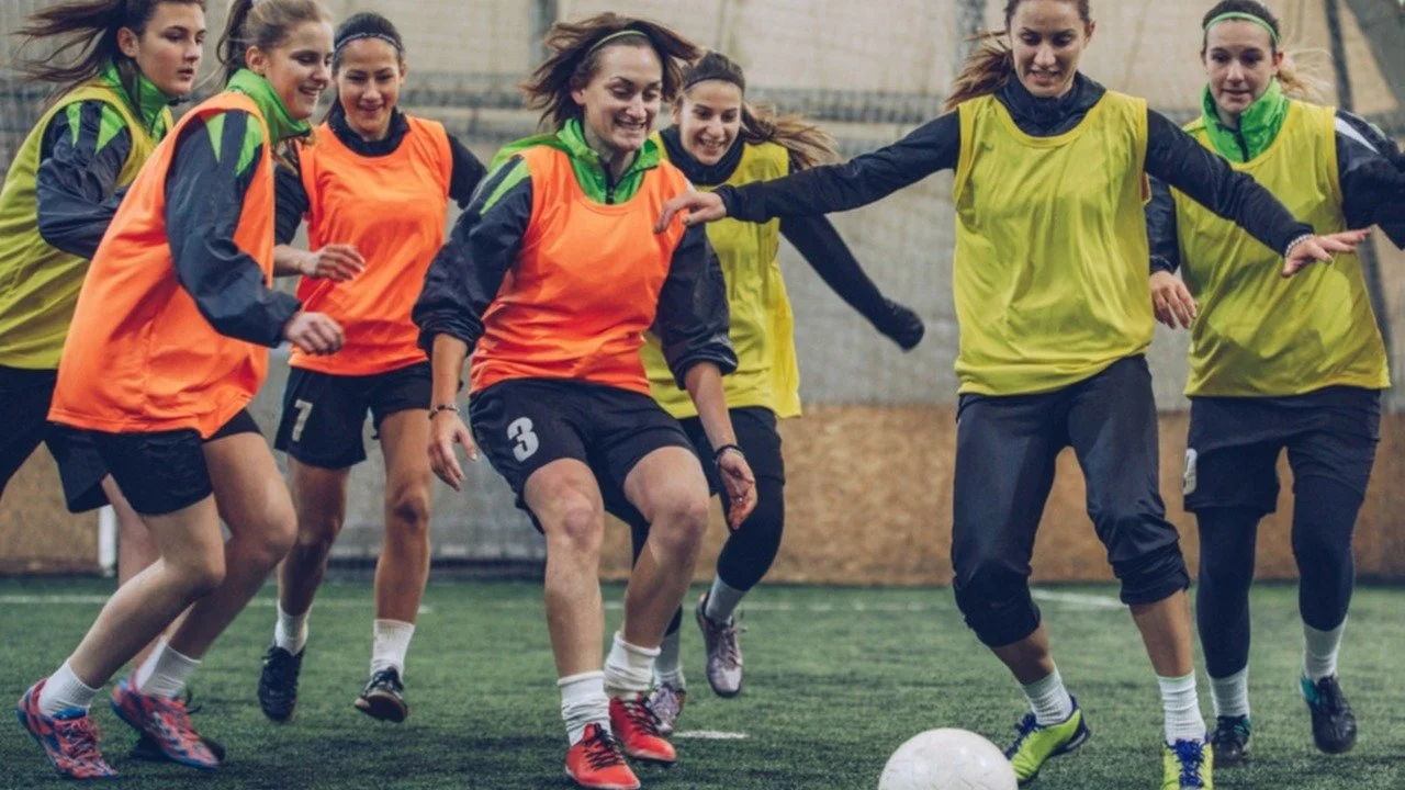 Group of women in orange and yellow bibs chasing a ball during a football session on an artificial pitch