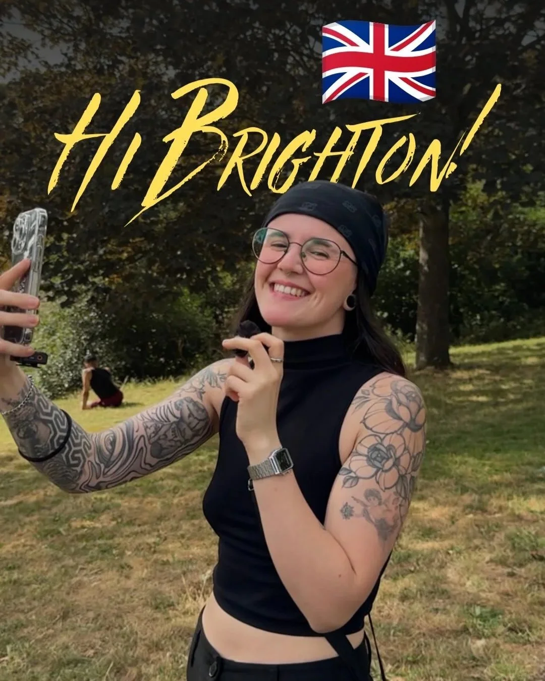 Spanish tattoo artist Copito smiling and filming a selfie video in a Brighton park, arms tattooed, with Hi Brighton written in gold above her