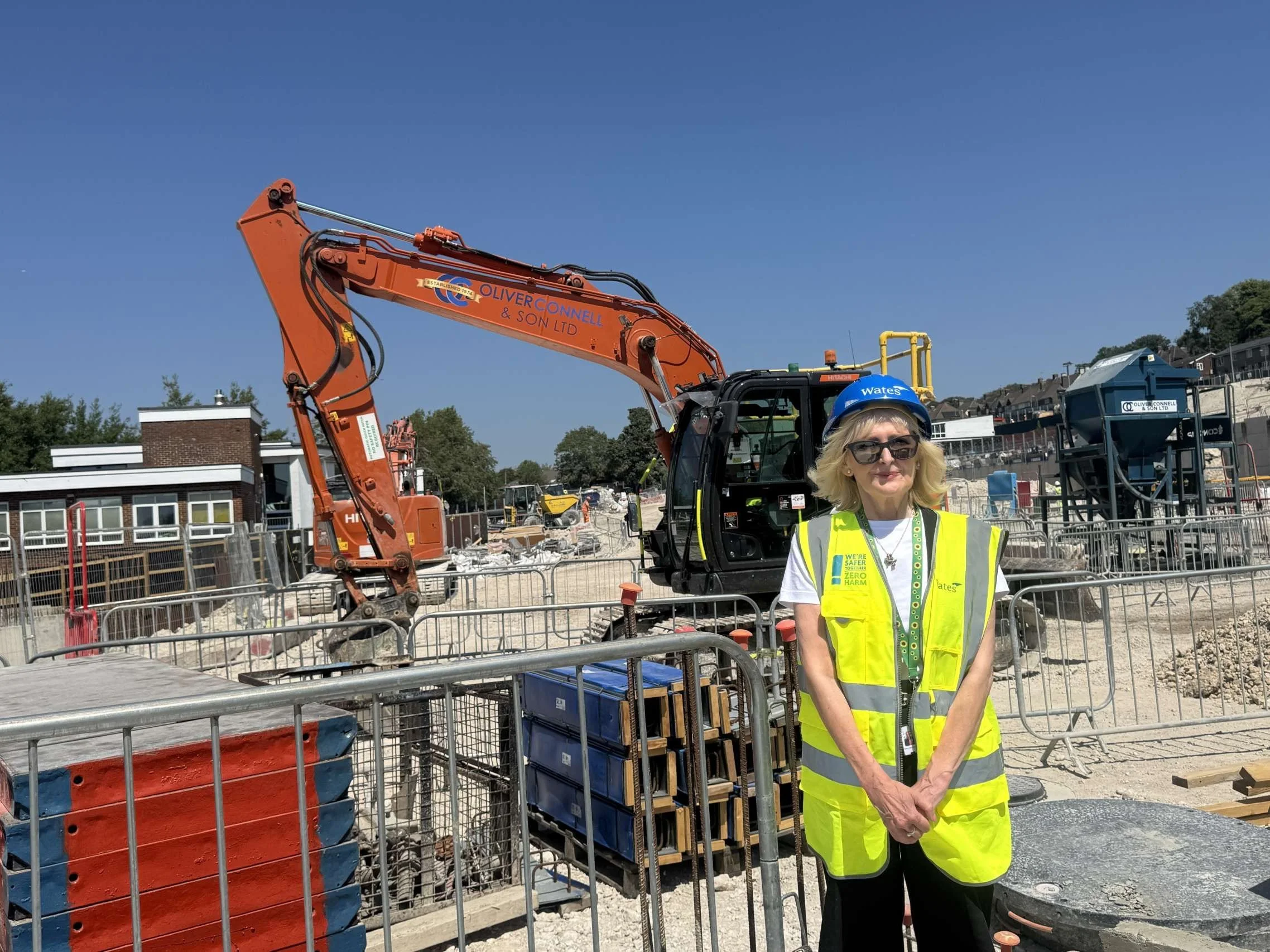 Councillor Gill Williams in a hi-vis vest and hard hat at the Moulsecoomb construction site with a digger behind her