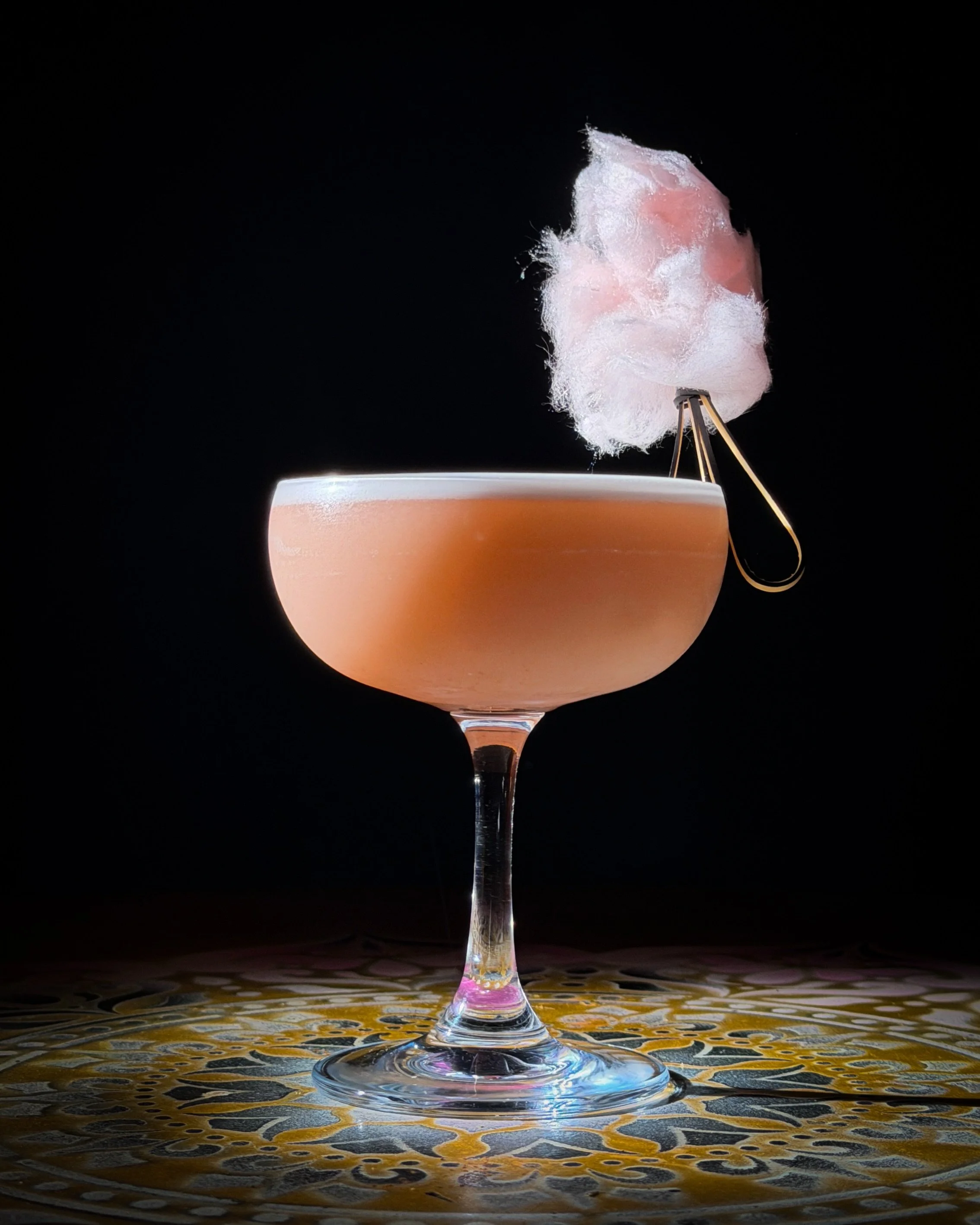 A pink cotton candy on a stick placed in a wine glass. The glass is on a circular patterned surface with a dark background.