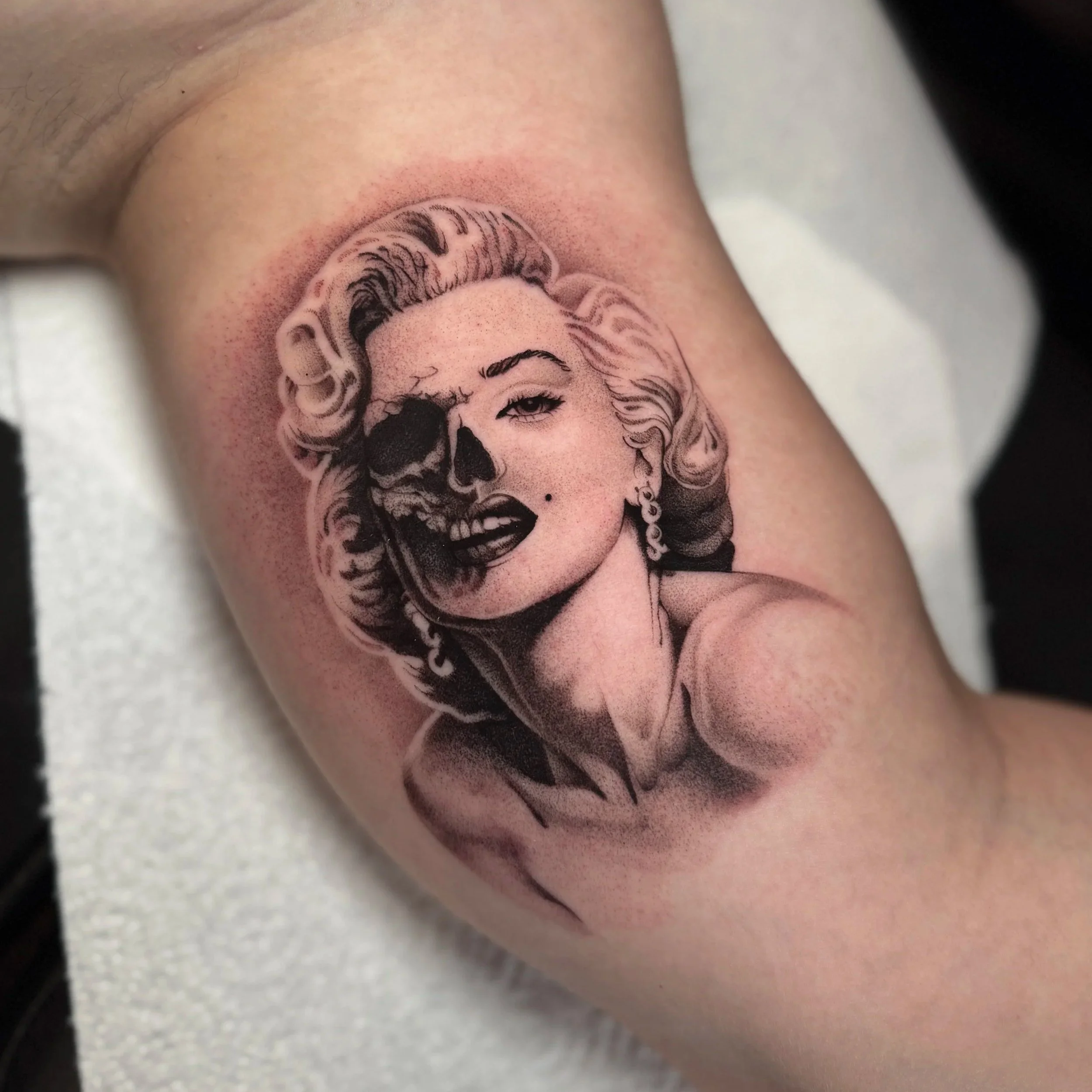 Black and grey inner-arm tattoo of Marilyn Monroe, one half her face and the other half a skull, in fine detailed shading
