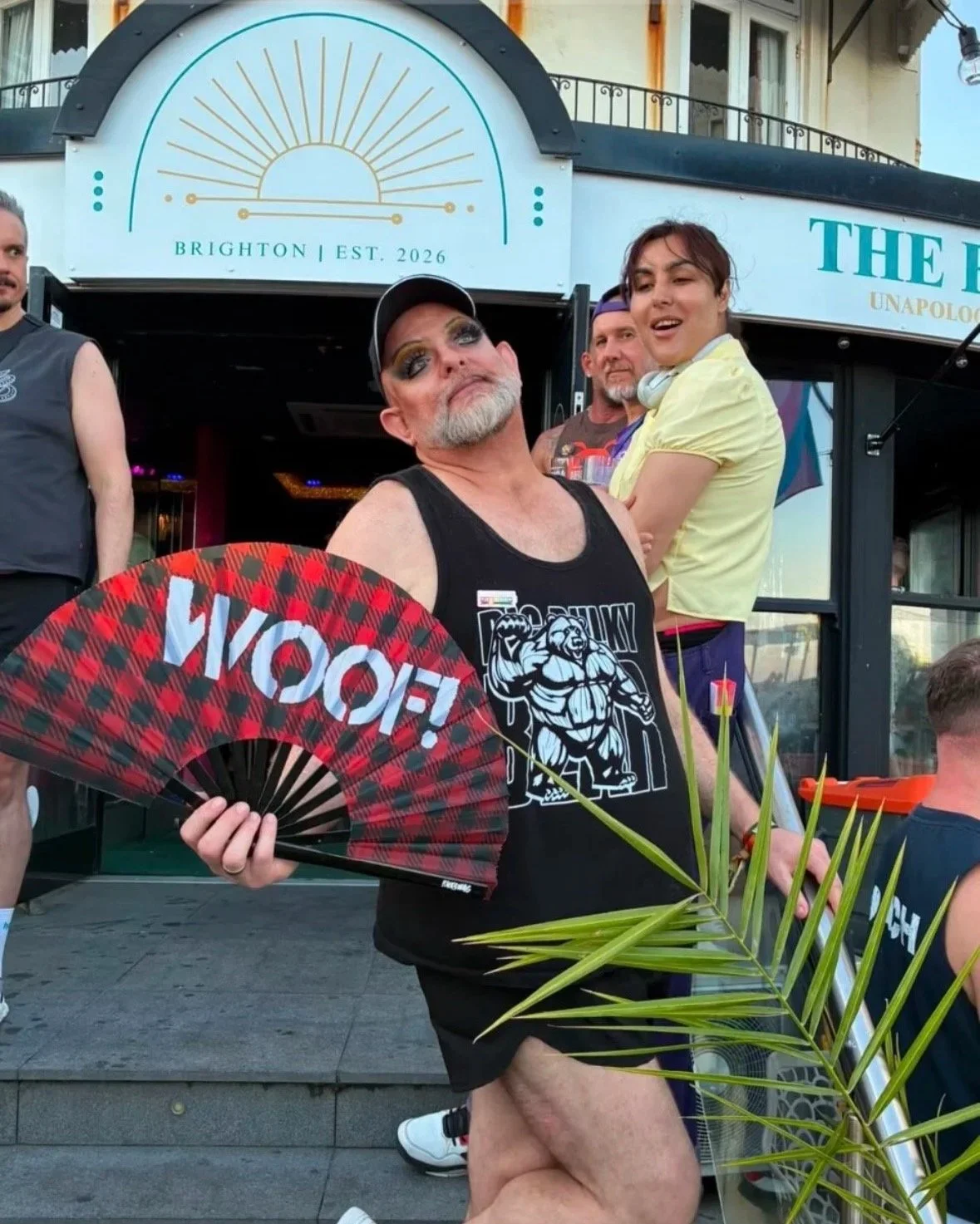 A drag performer poses outside The Birdcage on Marine Parade, Brighton, with a fan reading "Woof", other patrons in the background