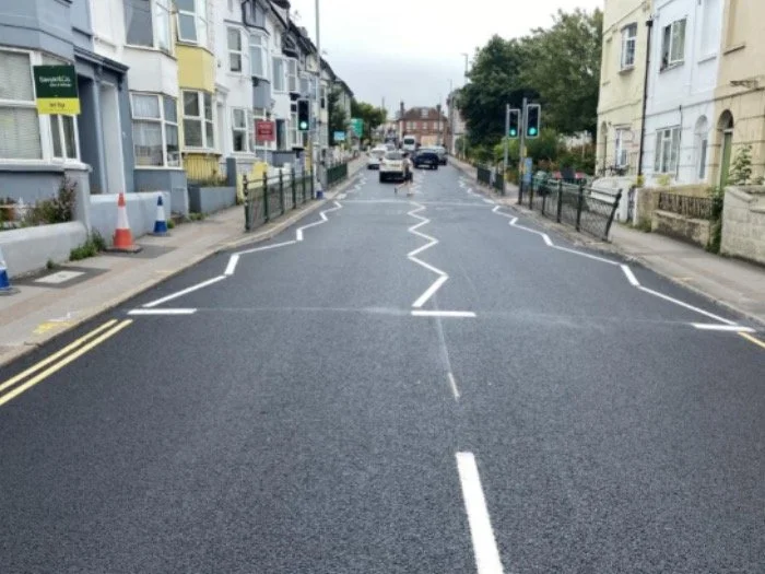 A resurfaced Brighton street with fresh white give-way and lane markings at a junction, terraced houses either side and traffic lights showing green