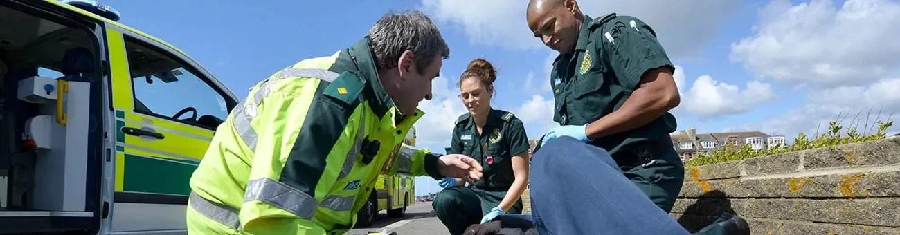 University of Brighton paramedic science students in high-visibility jackets and green uniforms treating a patient on the ground during an outdoor training exercise with an ambulance in the background
