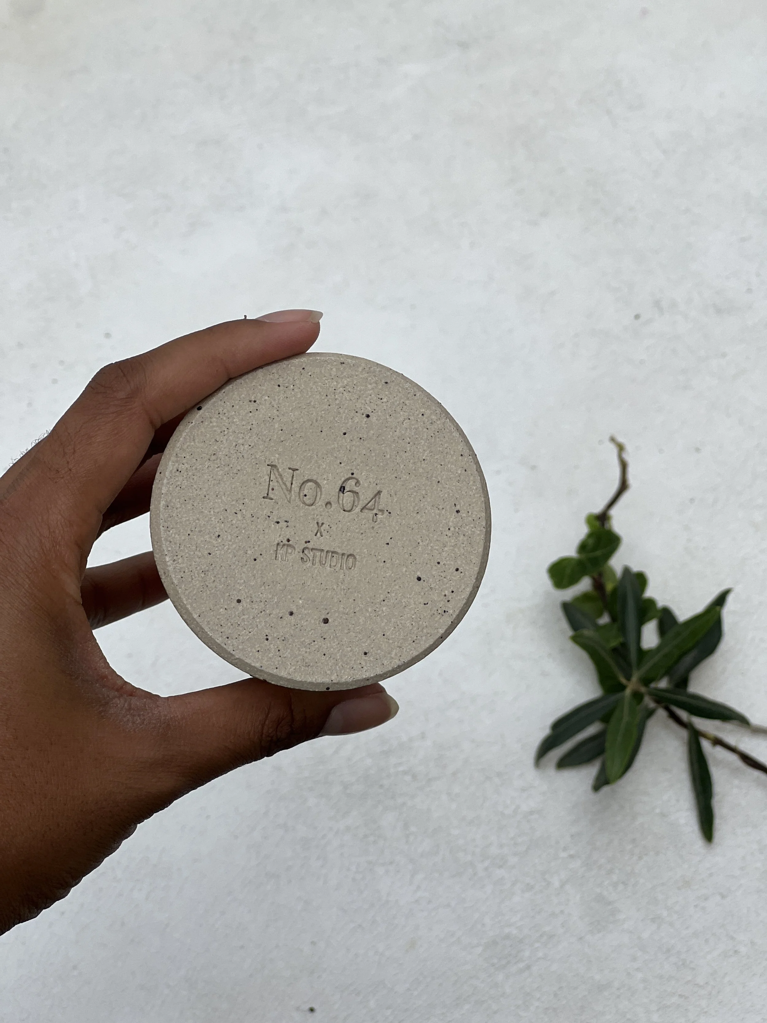 Hand holding a concrete-grey ceramic disc embossed with No.64 and KP Studio branding, with a fresh green plant stem beside it
