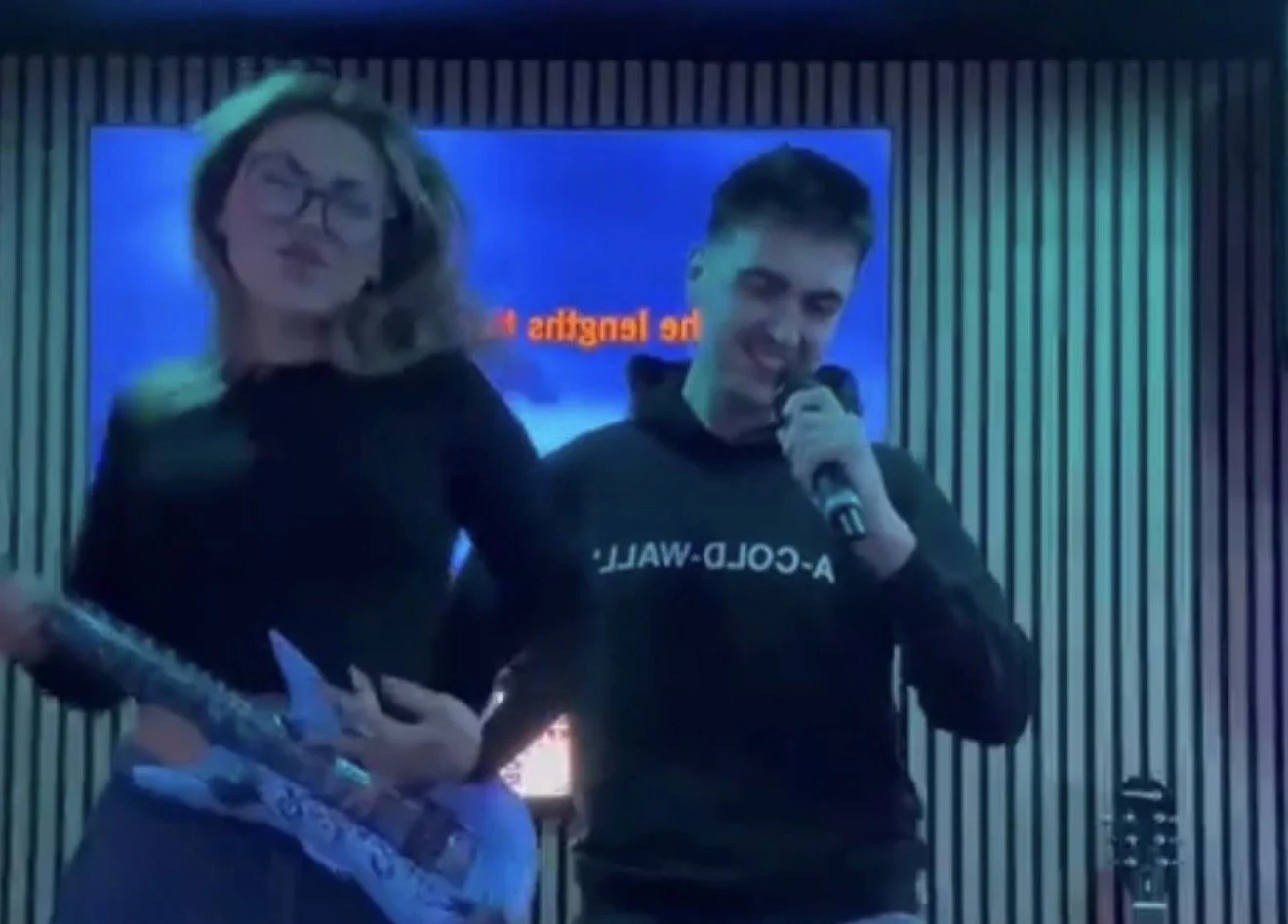 Two people singing karaoke at Ball Park Brighton Marina, one playing an inflatable guitar and the other singing into a microphone, in front of a lyrics screen and a slatted wood wall