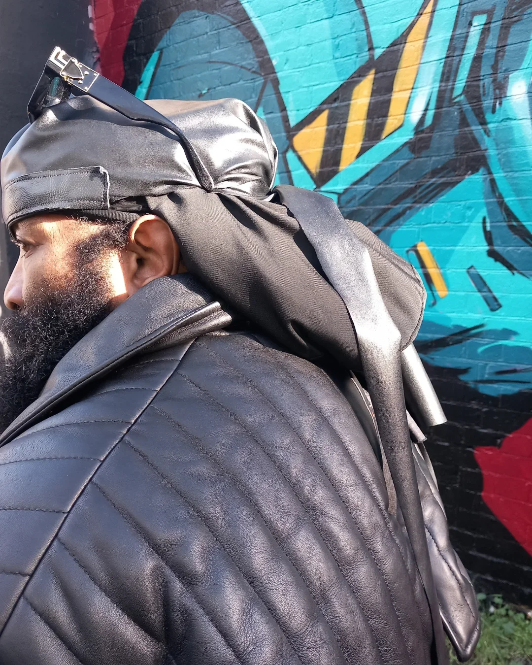 Charles Pirate Saddler in a black leather outfit and headwrap, side profile against a Brighton graffiti wall