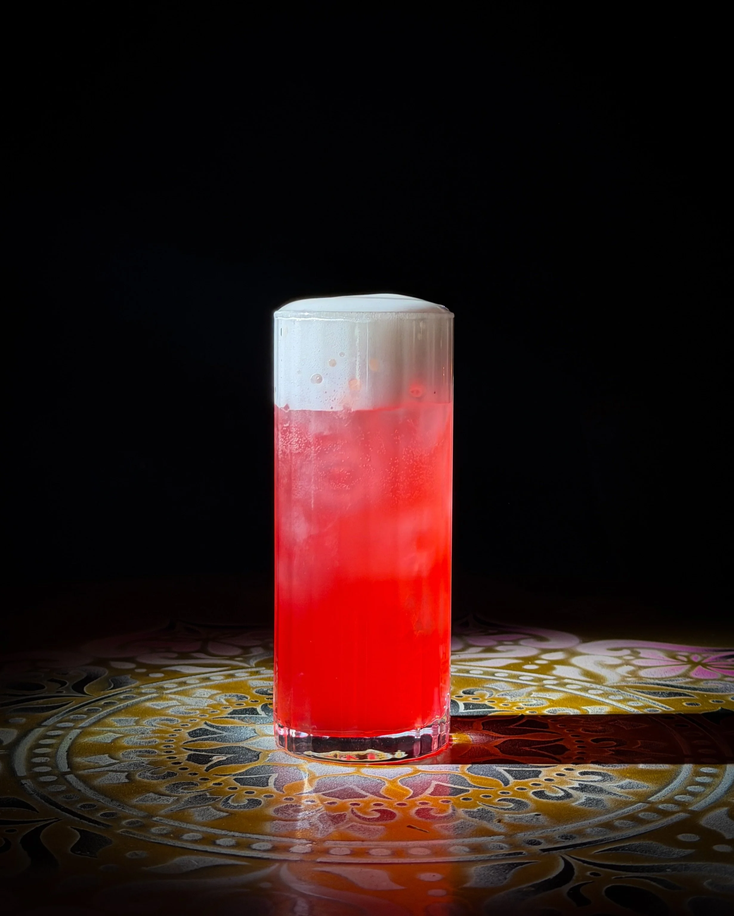 A tall glass with a shade of pink-red liquid and foam on top on a colorful, ornate table with a black background.