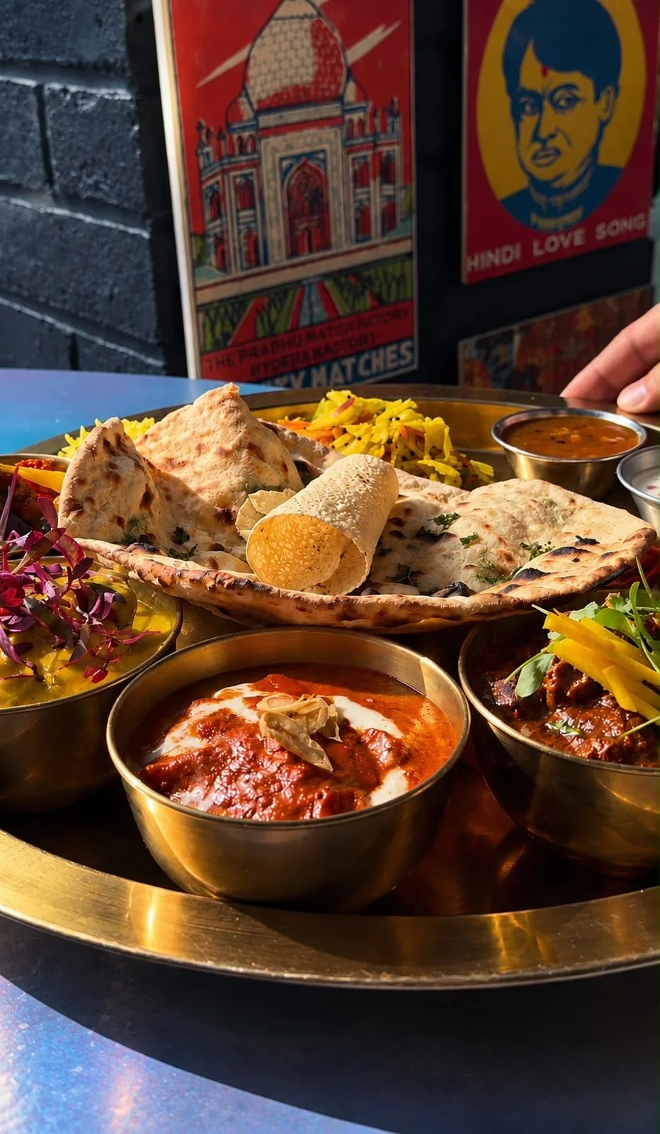 Side view of a brass thali tray at Indian Summer with bowls of curry, naan and a rolled papad in front of vintage Indian posters