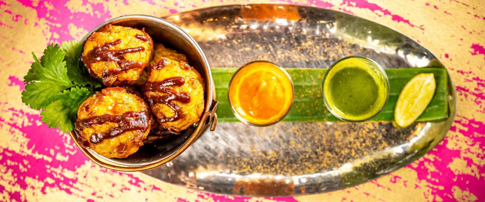 Top view of Samosa balls in a bowl with cilantro, served with two small glasses of chutney and a lemon wedge on a leaf, on a metal tray with pink and yellow patterned cloth underneath.