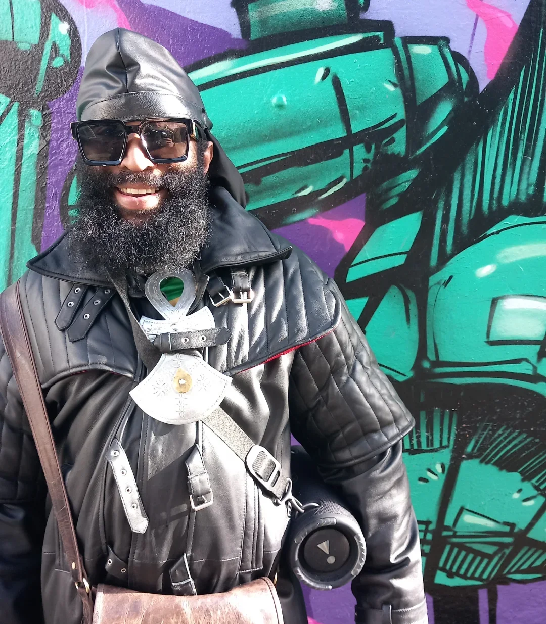 Charles "Pirate" Saddler smiling in sunglasses, a black leather jacket with metal shoulder pauldrons and an ankh pendant, standing in front of a green and purple graffiti mural