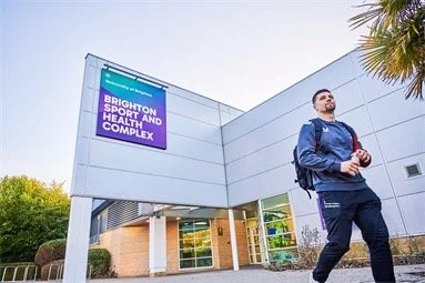 Modern University of Brighton complex building exterior with illuminated purple sign and a person in paramedic uniform outside