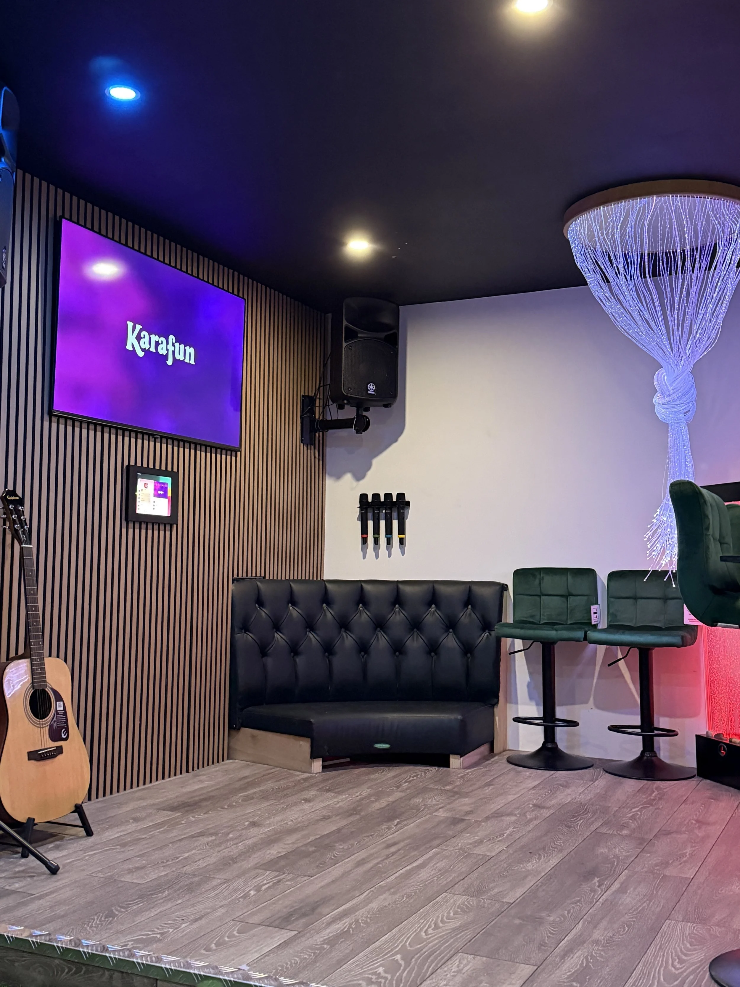 The VAR Room karaoke suite at Ball Park Brighton Marina with a Karafun screen, wireless microphones on the wall, a black leather corner booth, green velvet bar stools, an acoustic guitar and a fibre optic light feature