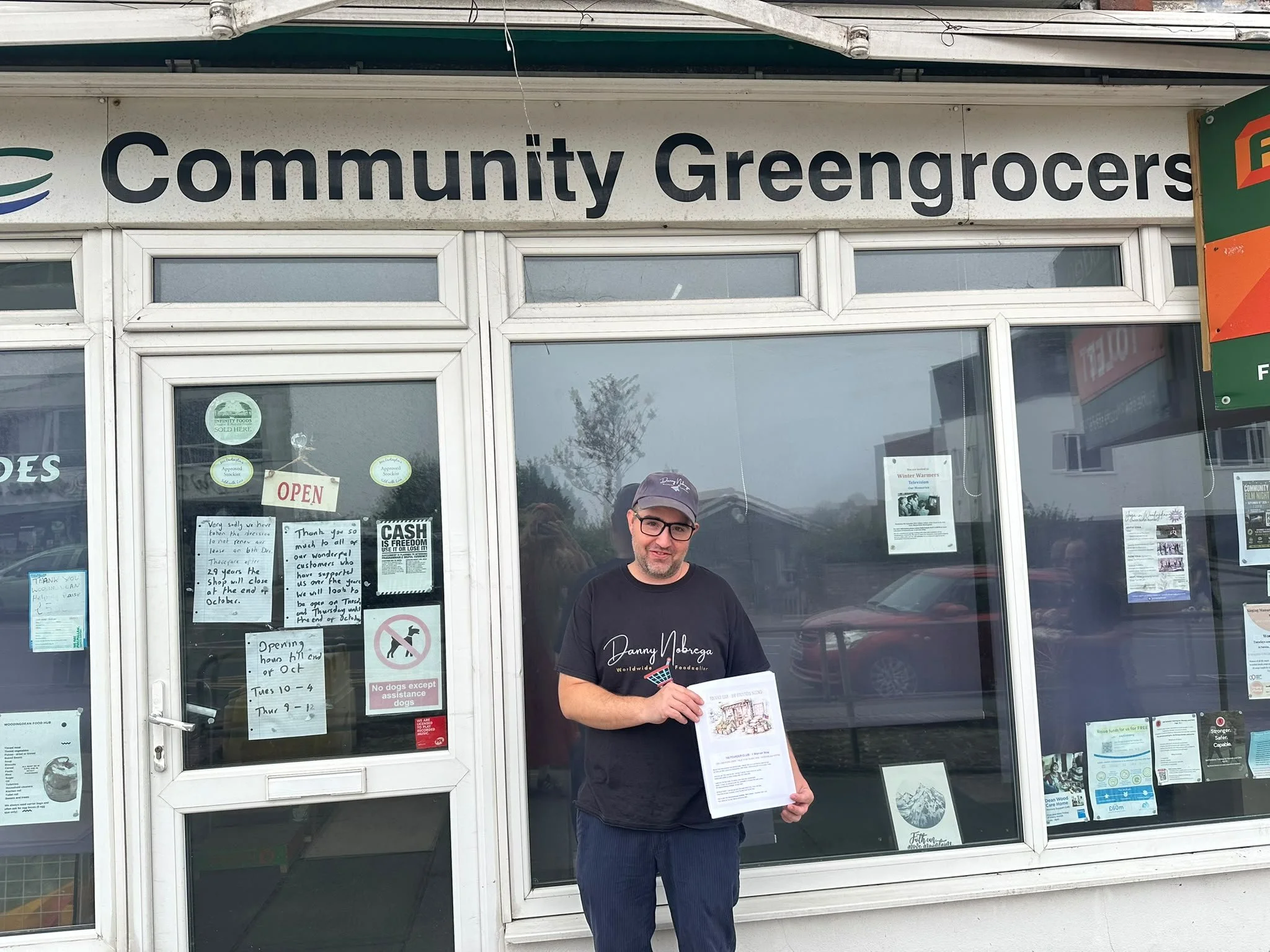 Danny Nobrega standing outside Woodingdean Community Greengrocers under the shop's sign, holding his Founder Club flyer, with closure notices visible in the window behind him
