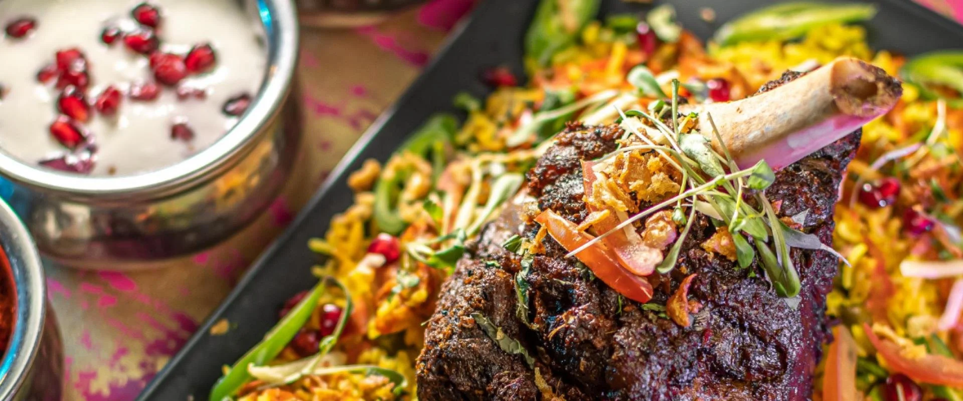 Close-up of a cooked beef rib served on a bed of colorful vegetables, garnished with microgreens. Pomegranate seeds are visible in a bowl nearby.