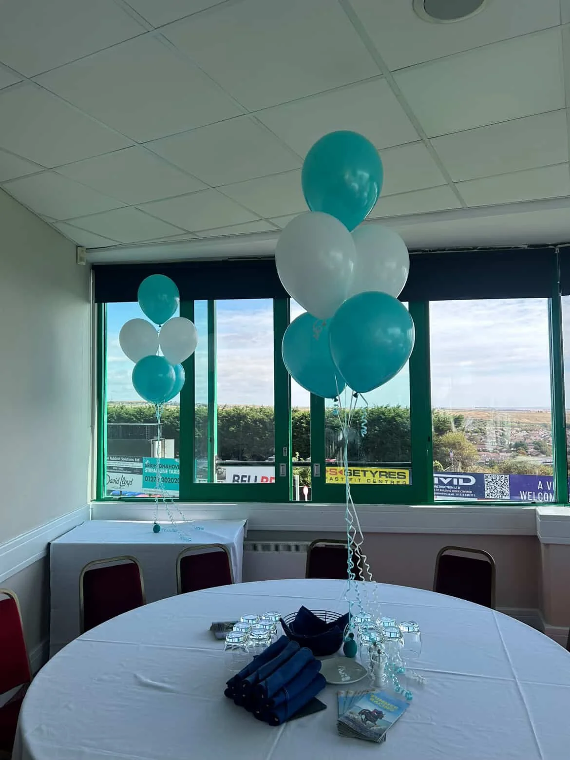 Table set with glasses and racecards beneath teal and white balloons at Streamline Taxis' Martlets Hospice evening