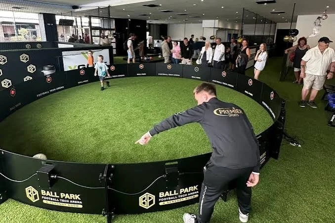 A coach running a football session in the ring arena at Ball Park Brighton Marina as a crowd watches