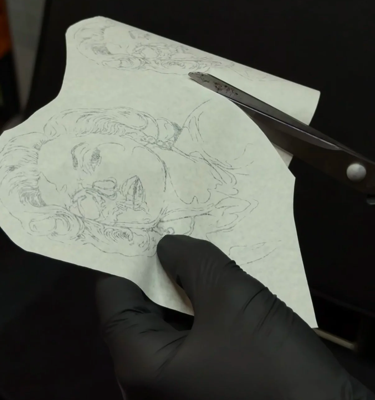 A gloved hand holding the paper stencil of the Marilyn Monroe design, cut to shape with scissors, before tattooing