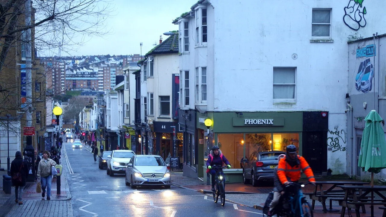 Trafalgar Street in Brighton at dusk, with cyclists and cars on a wet road and shops and cafes along the side of the street