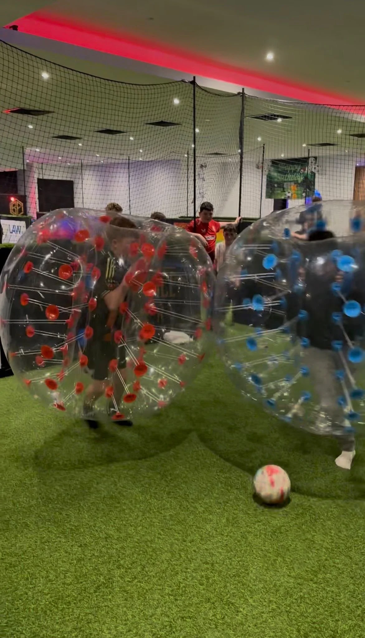 Two children playing bubble football inside clear inflatable Zorb balls with red and blue grips on the artificial turf at Ball Park Brighton Marina, with a football on the pitch and a Happy Birthday banner behind them