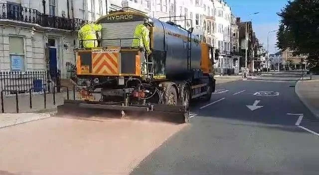 A Hydrog microasphalt treatment vehicle laying a fresh road surface on a residential Brighton street, with two workers on the rear platform